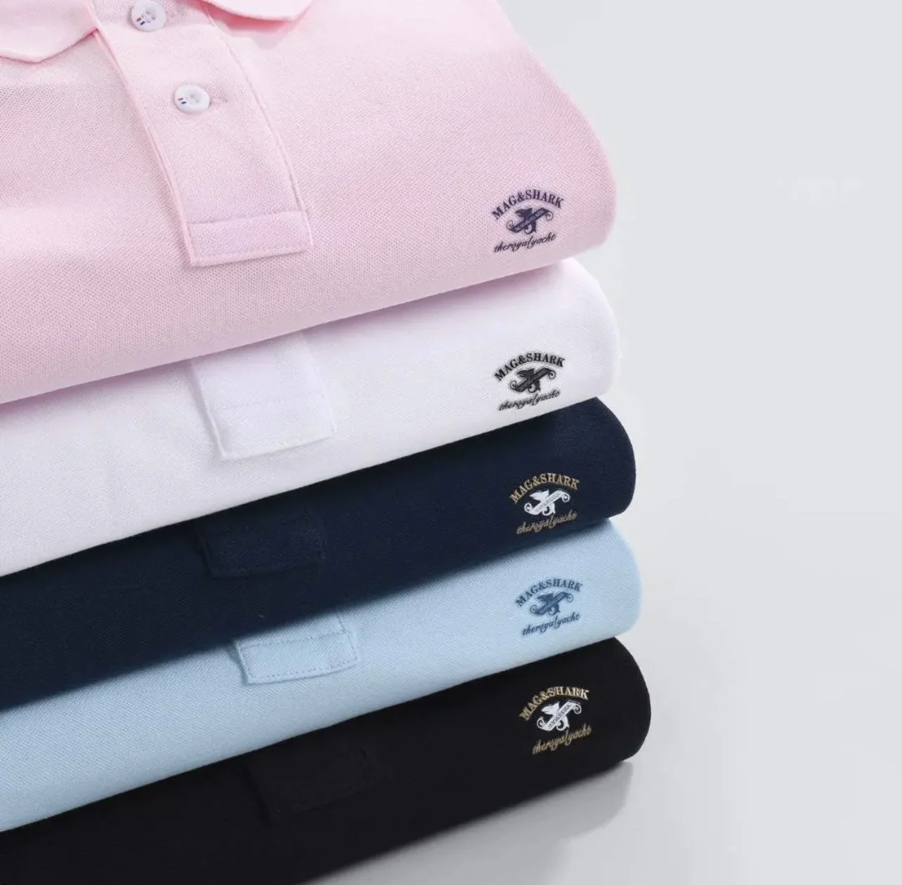 A stack of folded polo shirts in pink, white, navy blue, light blue, and black with embroidered logo "MAG & SHARK".
