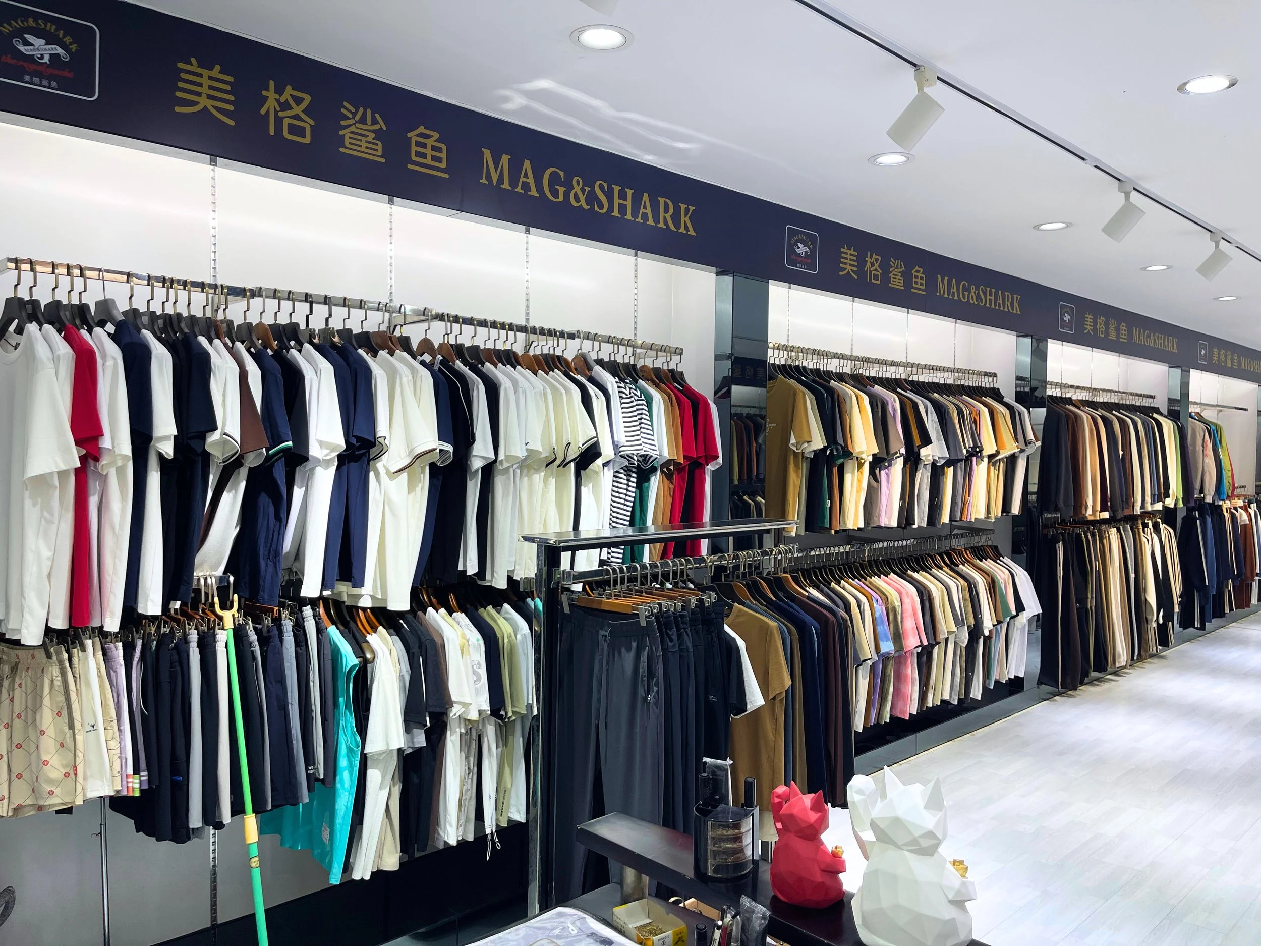 A clothing showroom with racks of shirts and pants, featuring a sign that reads 'MAG&SHARK' in English characters.