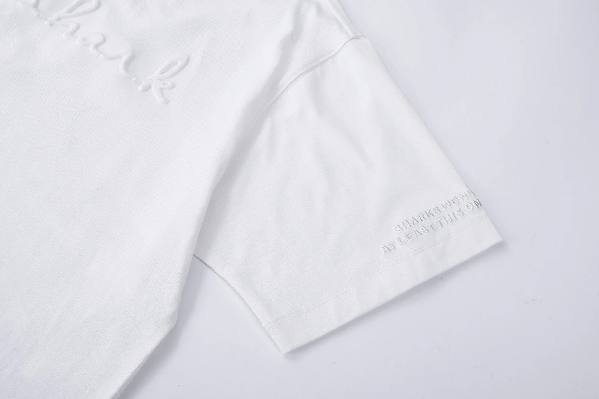 Close-up of a white t-shirt with embroidered text and a graphic design, with some embossed text on the sleeve.