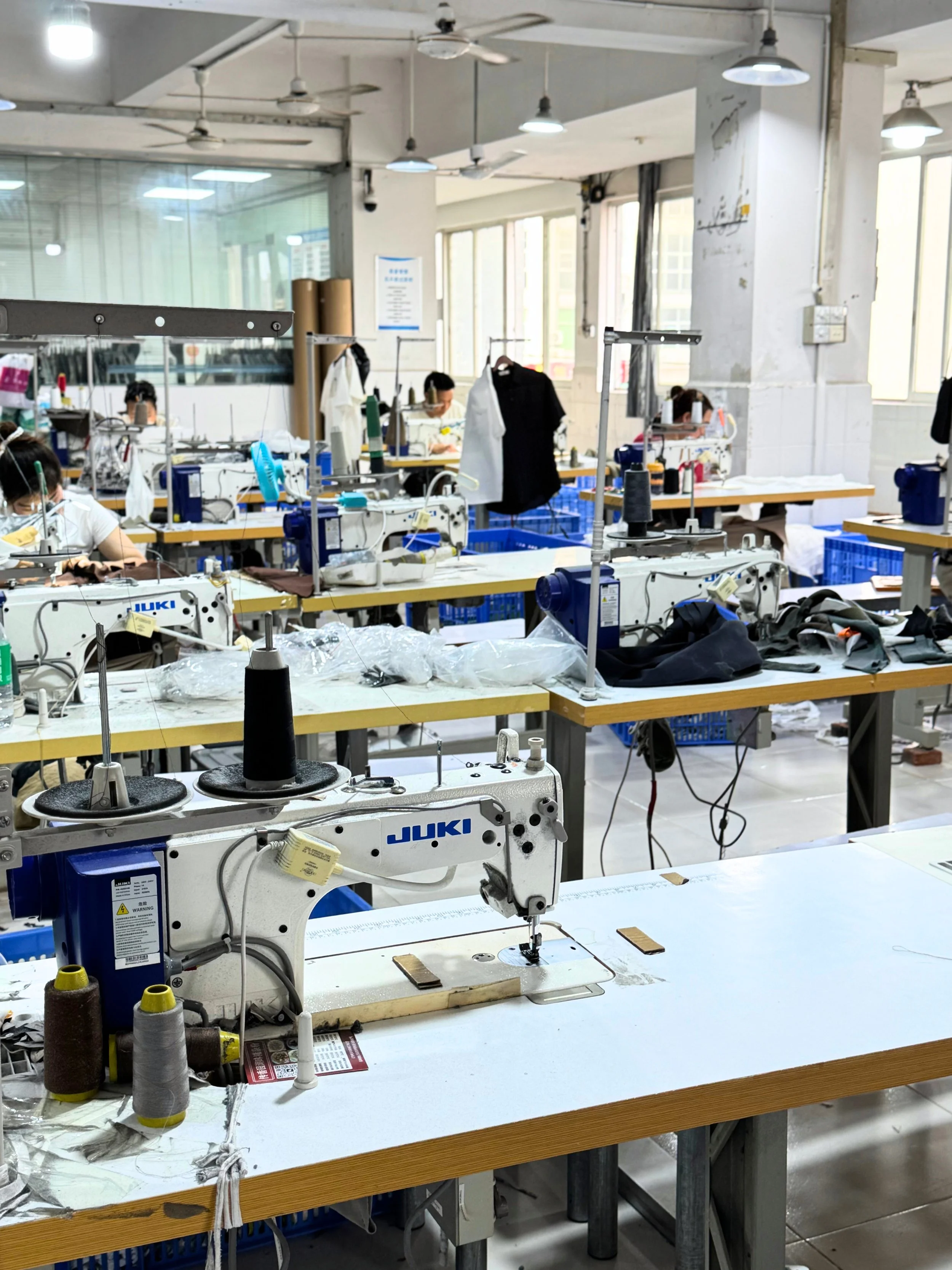 A sewing workshop with multiple workstations, industrial sewing machines, fabrics, and workers wearing face masks working on garments.