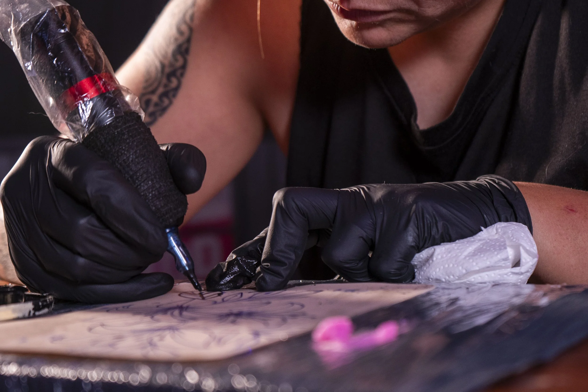 Une personne tatouée et portant des gants noirs utilise un stylo pour dessiner ou tatouer sur une surface avec une concentration intense.