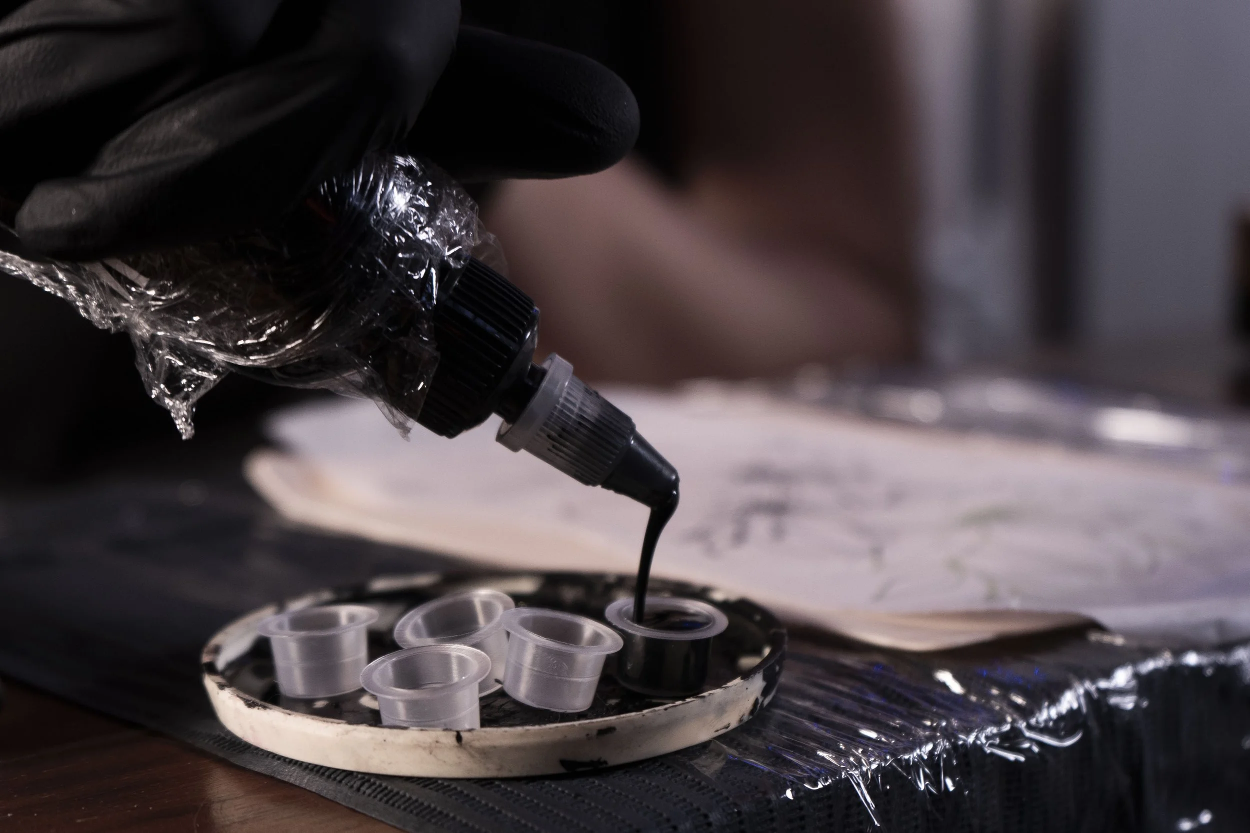 Main portant un gant noir verse de l'encre de tatouage dans un petit récipient à côté d'autres récipients vides sur une surface recouverte de film plastique.