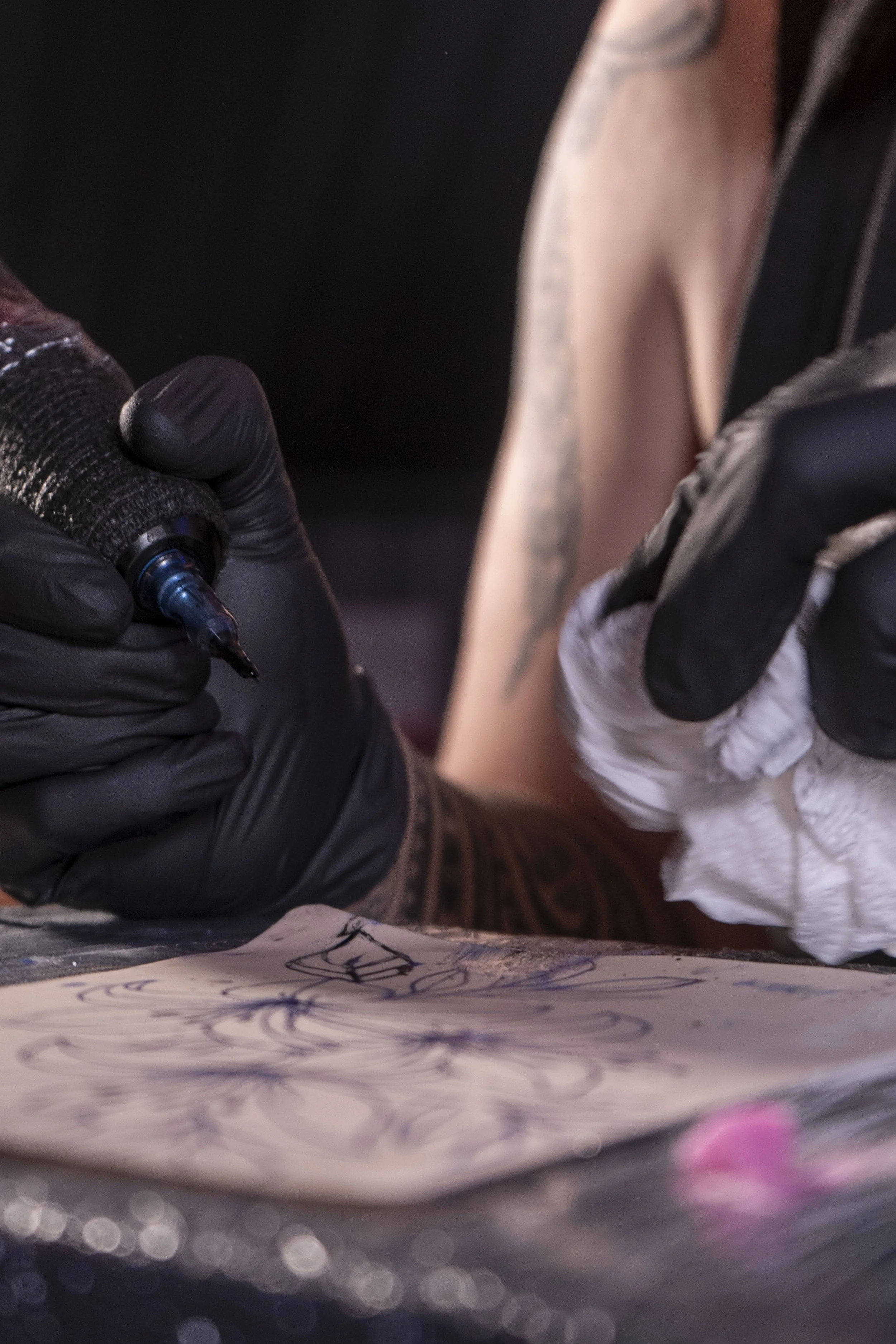 Une personne tatouée en train de faire un tatouage sur un bras, portant des gants noirs et utilisant une machine à tatouer.