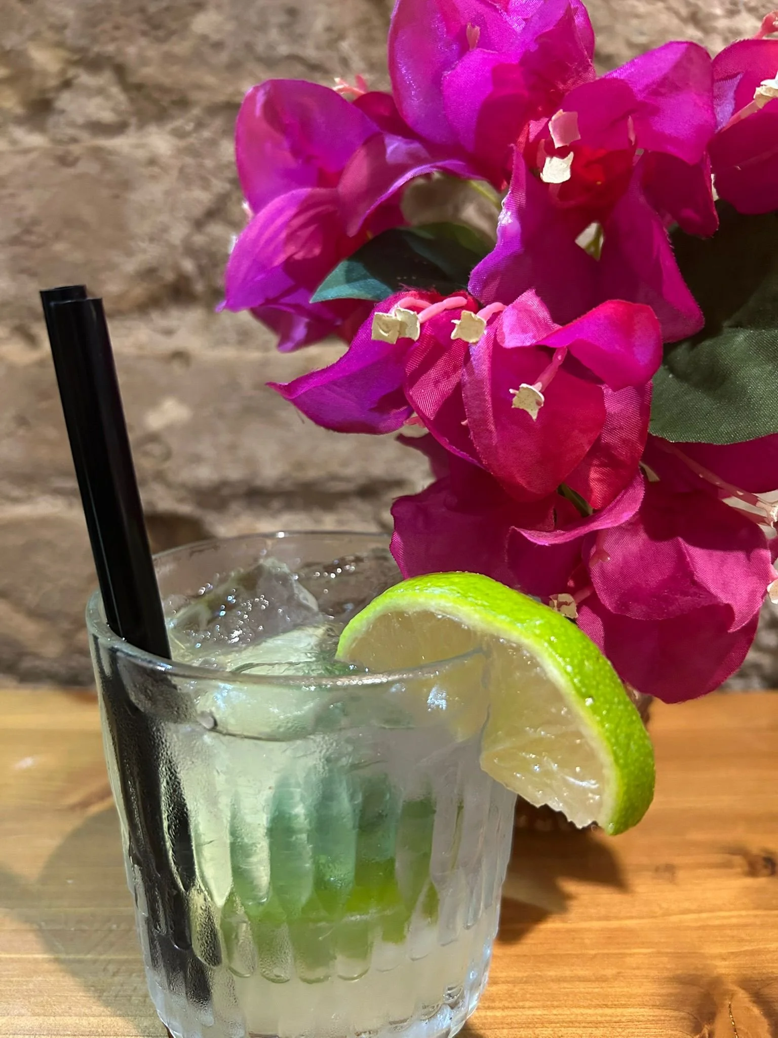 Un bicchiere di cocktail con lime e ghiaccio, decorato con una fetta di lime, sul tavolo di legno con un fiore di bougainvillea rosa sullo sfondo.