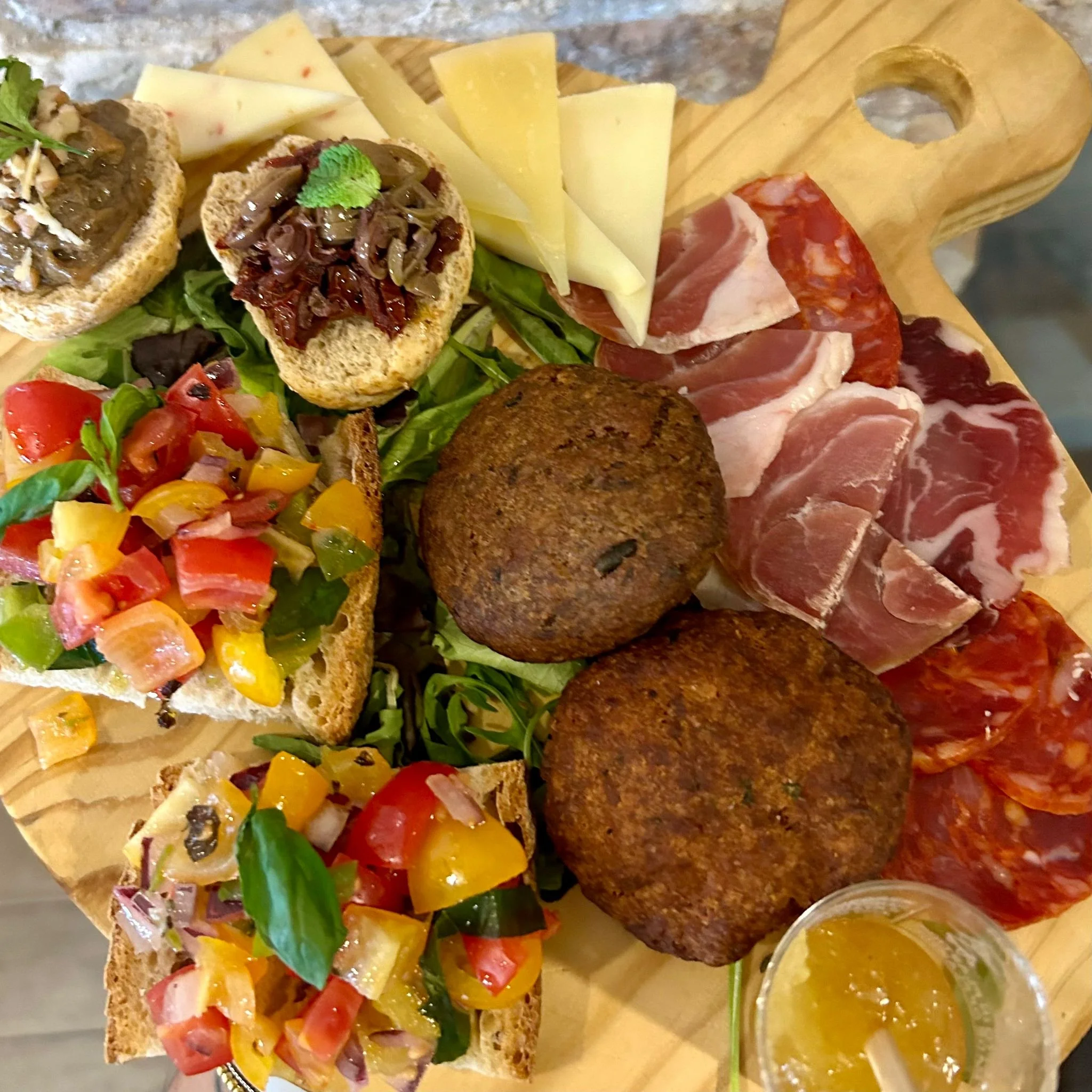 Un tagliere con affettati, formaggi, crostini con verdure, polpette e un bicchiere di marmellata.