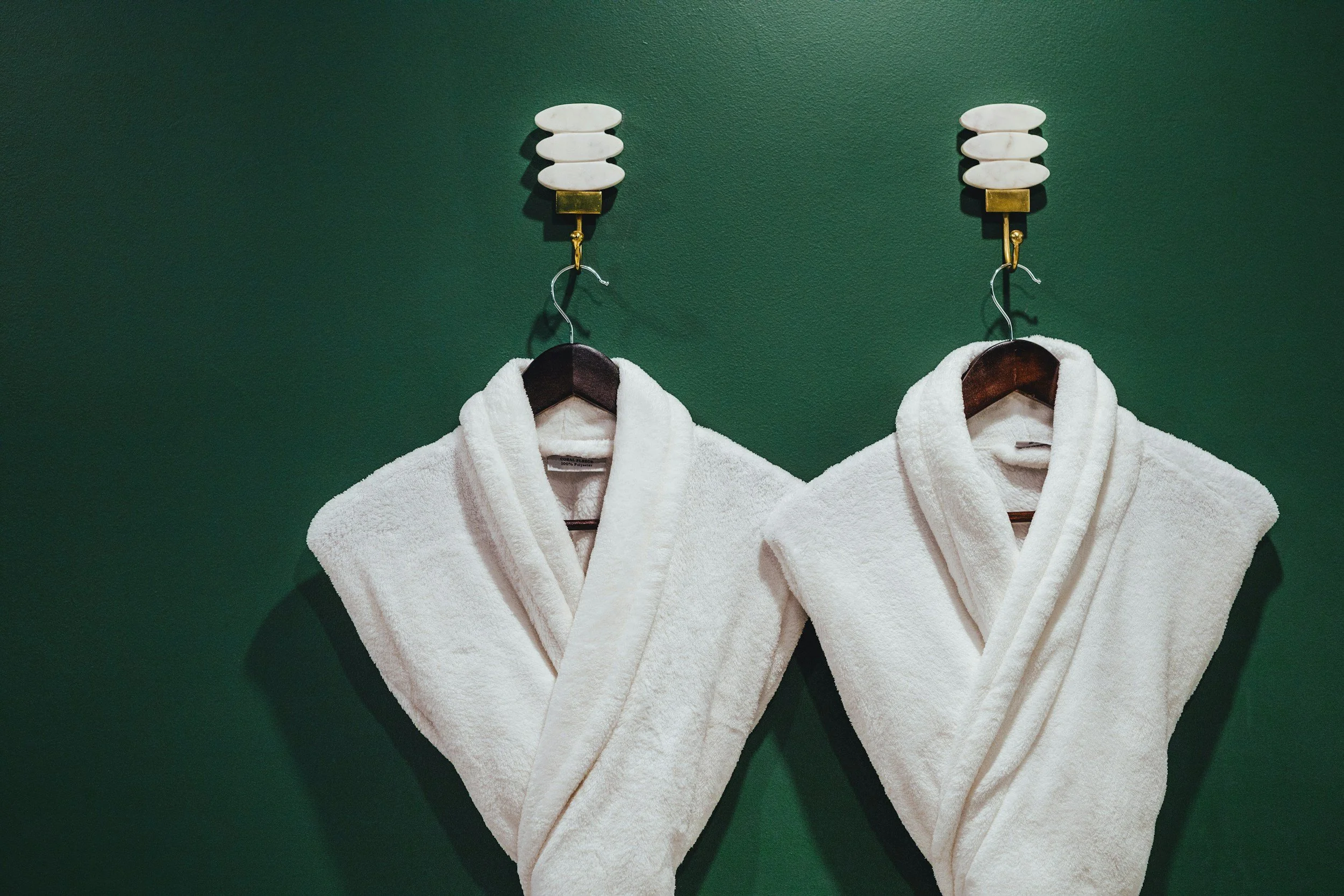 Two white bathrobes hanging on wooden hangers on a dark green wall, with decorative wall-mounted light fixtures above.