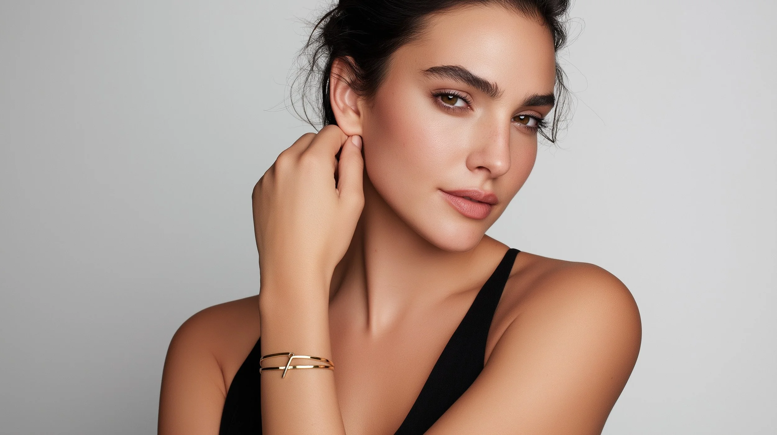 A woman with dark hair and light skin wearing a black tank top and gold jewelry posing against a plain background.