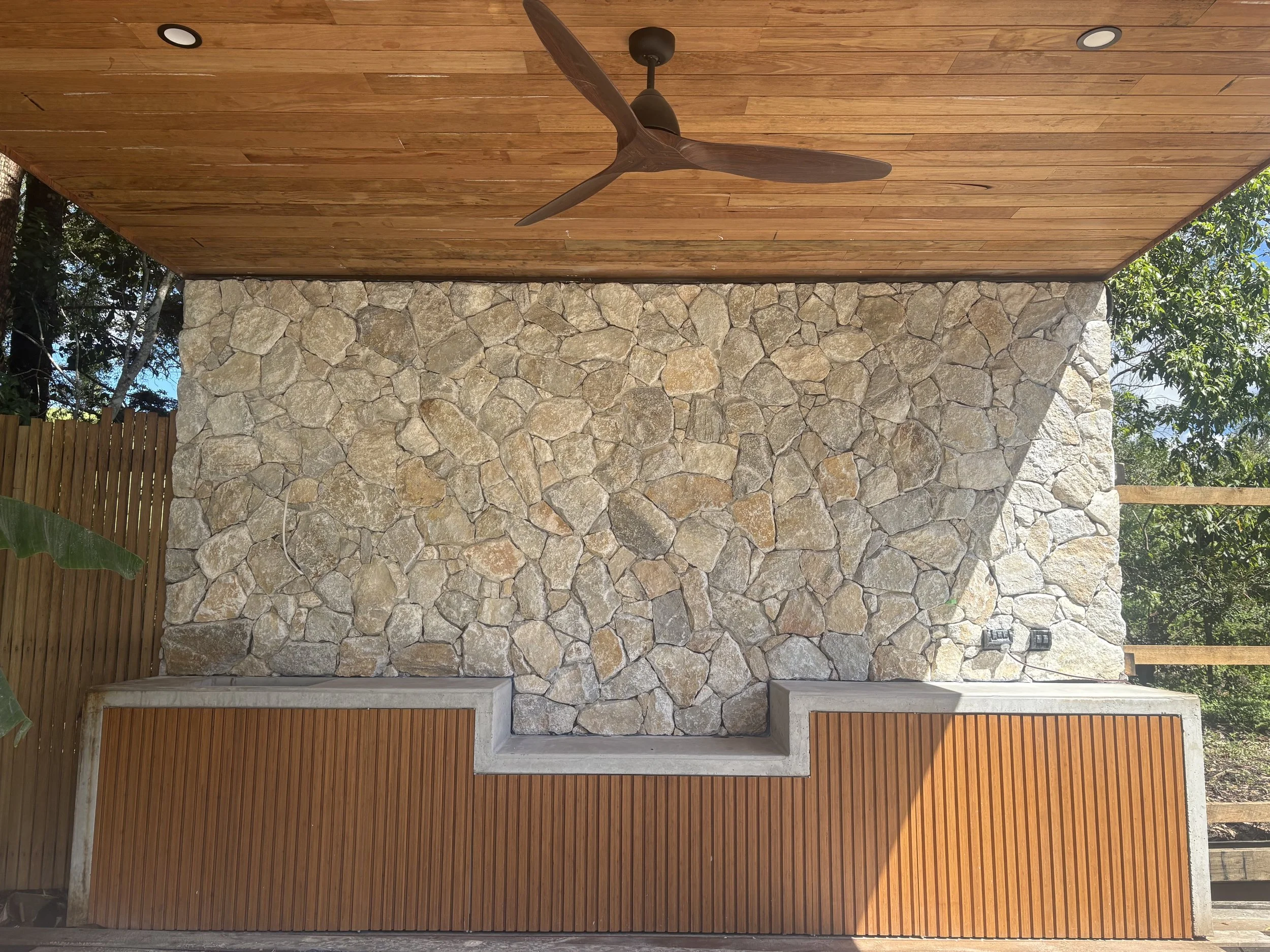 Outdoor ceiling with wooden panels, a ceiling fan, and recessed lighting. A stone wall with irregularly shaped stones and a concrete base with wooden slats below. Electrical outlets are visible on the right side of the stone wall.