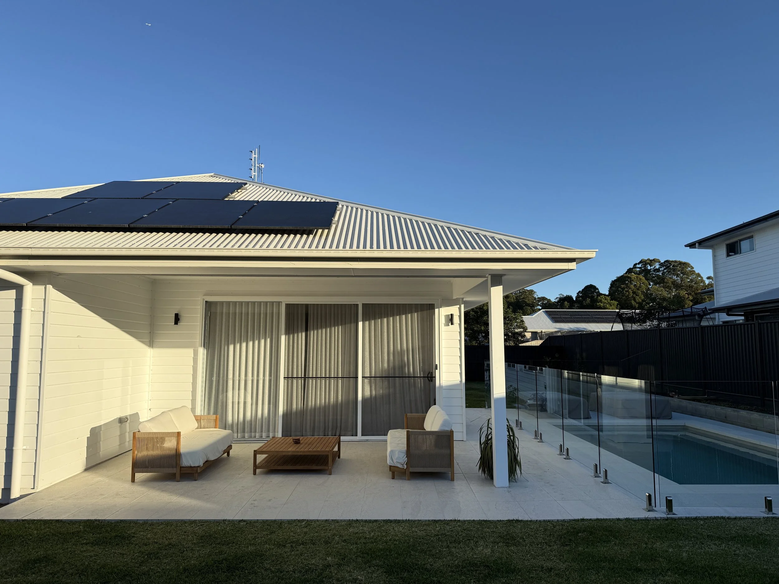 Modern backyard with tiled patio, outdoor seating area, glass pool fencing, and a swimming pool, under a clear blue sky.