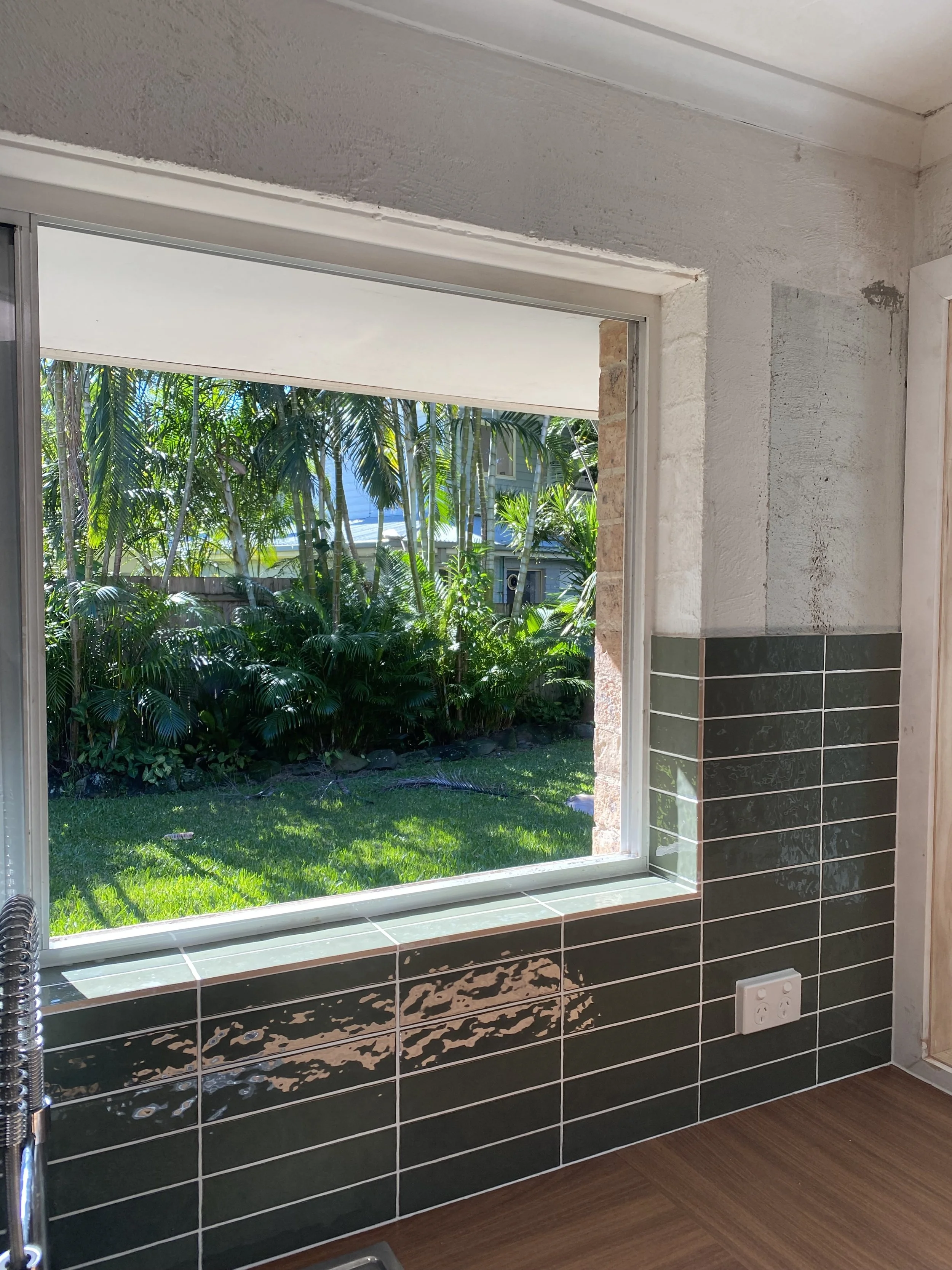View of lush green tropical plants and trees through a large window in a room with partially tiled walls and a power outlet on the wall.