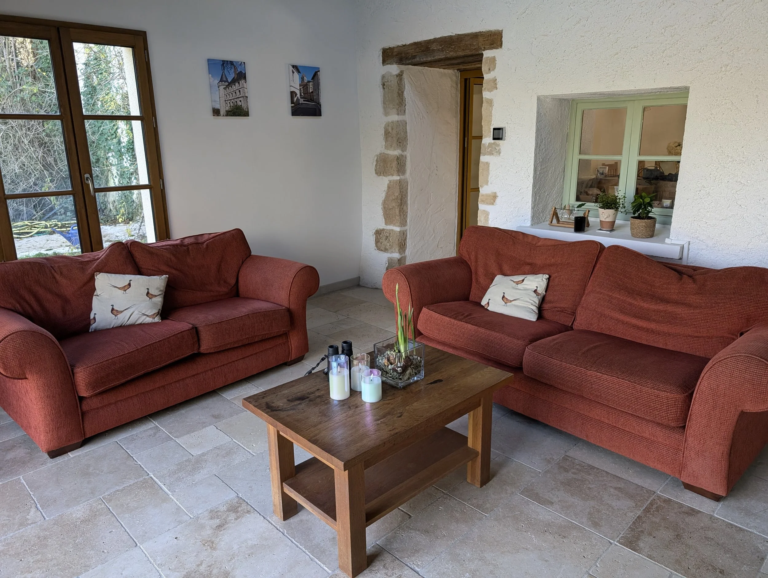 Living room with two red sofas, a wooden coffee table with candles and a plant, large window with a garden view, and small picture frames on the wall.