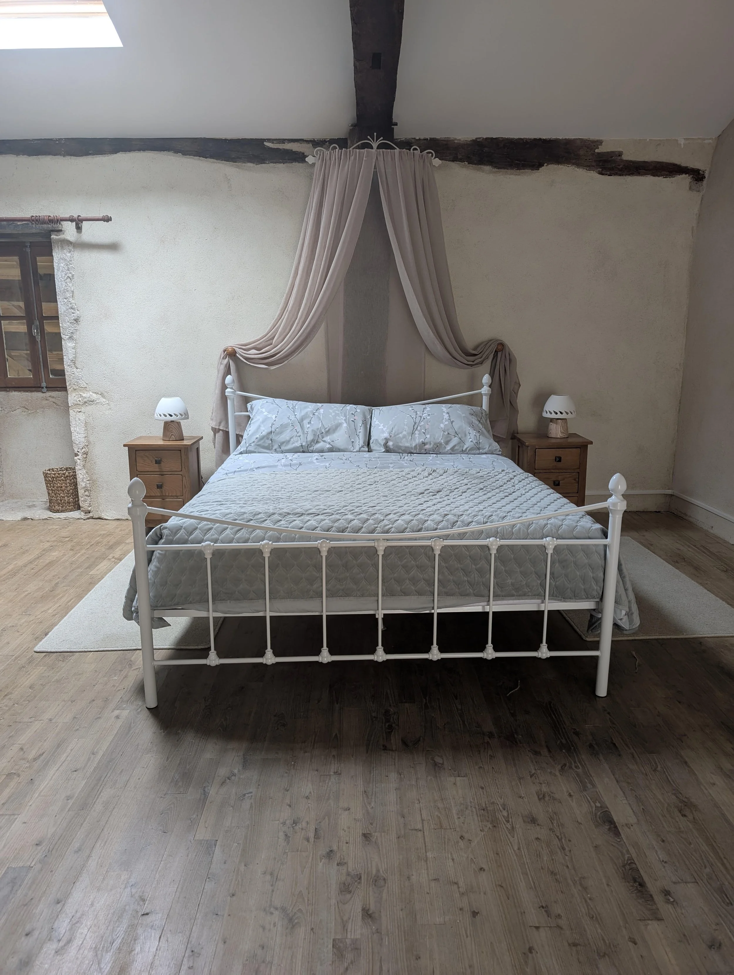 A bedroom with a white metal bed frame, light-colored bedding, two wooden nightstands with lamps, and a beige curtain draped above the bed in a rustic room with exposed wooden beams and a skylight.
