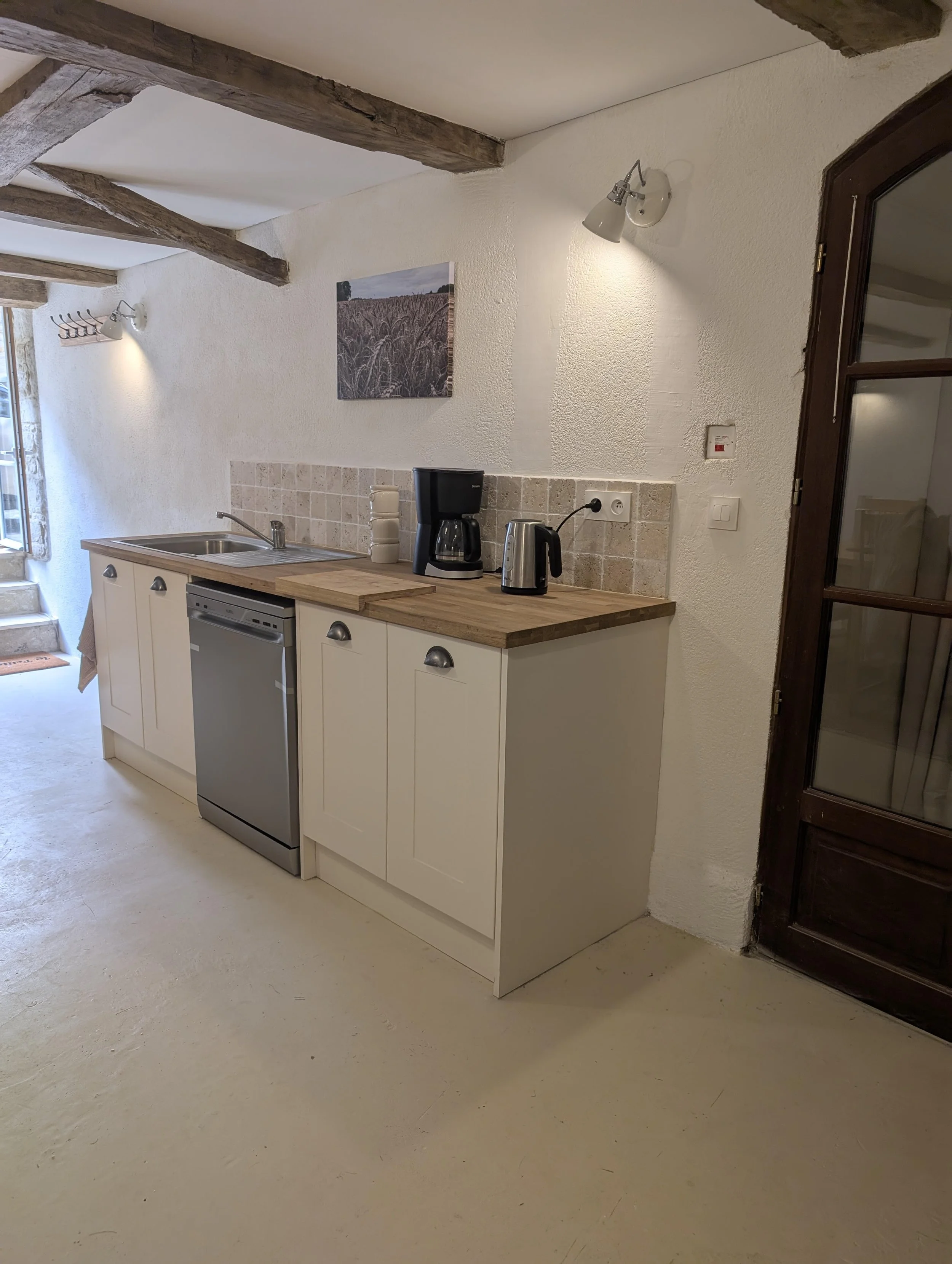 Small kitchen with beige cabinets, wooden countertop, coffee maker, electric kettle, and tile backsplash, in a room with exposed wooden beams and a window.