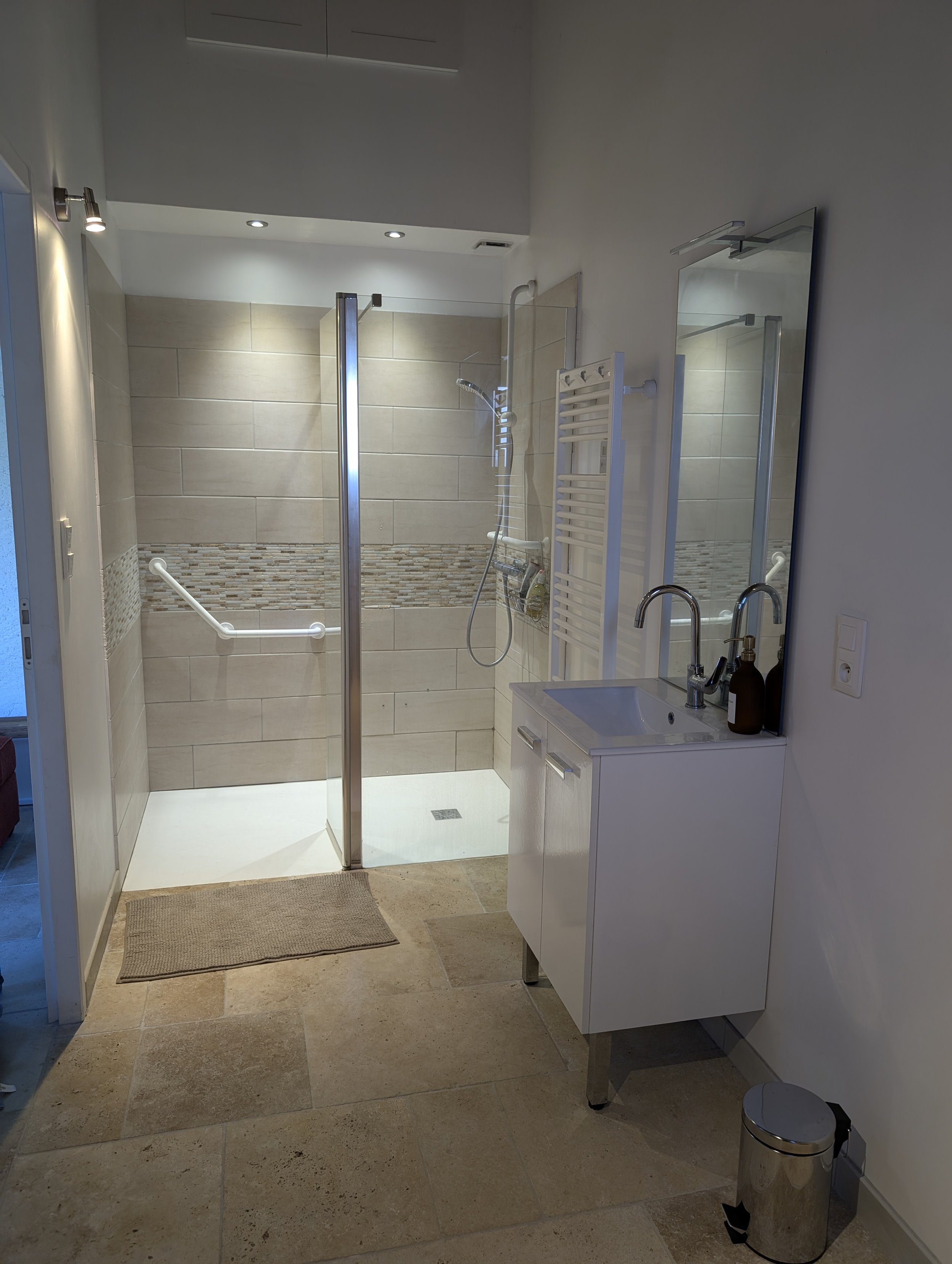 Bathroom with walk-in shower, small white sink cabinet, large mirror, and beige tiled floor.