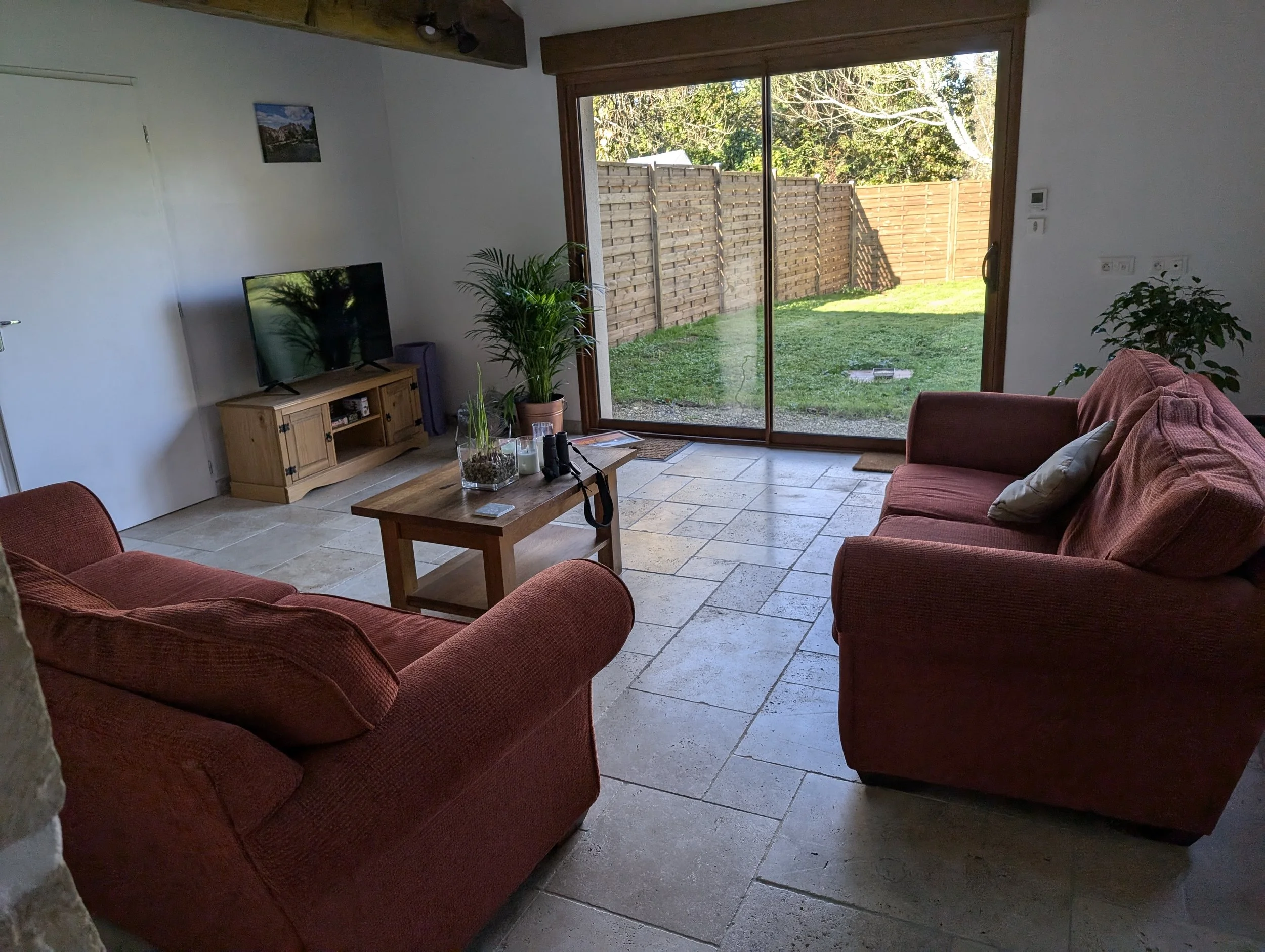 Living room with two red couches, a wooden coffee table, a TV on a wooden stand, plants, and large glass sliding doors leading to a backyard with a grassy area and a wooden fence.
