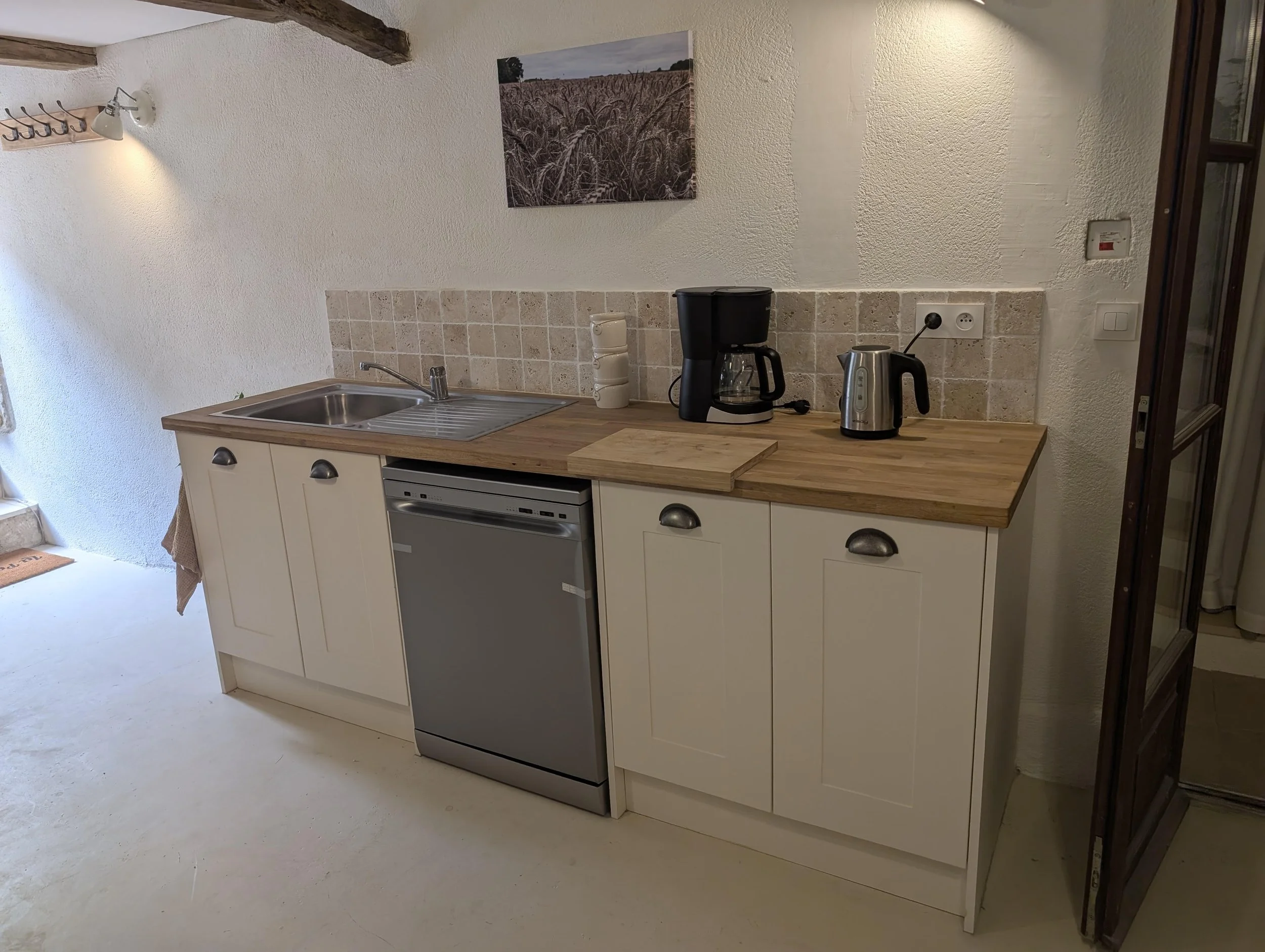 Kitchen workspace with white cabinets, a wooden countertop, a sink, a coffee maker, a kettle, and a small dishwasher, with a picture of a field on the wall.
