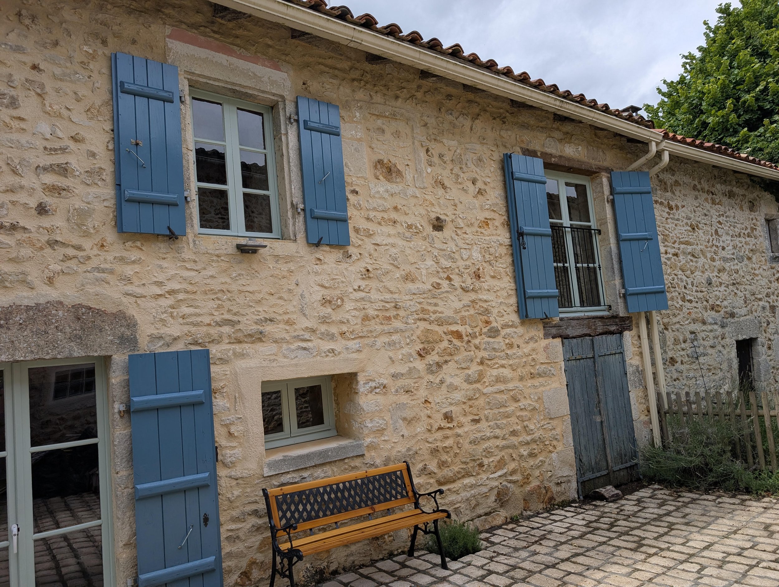 Stone house with blue shutters, multiple windows, a wooden door, and a bench outside on cobblestone pavement.