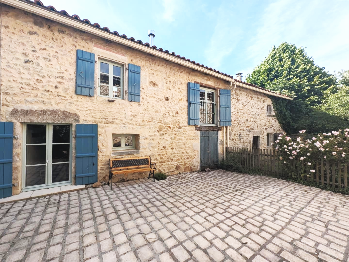 Stone house with blue shutters, cobblestone patio, wooden bench, and blooming pink flowers, under a clear blue sky.