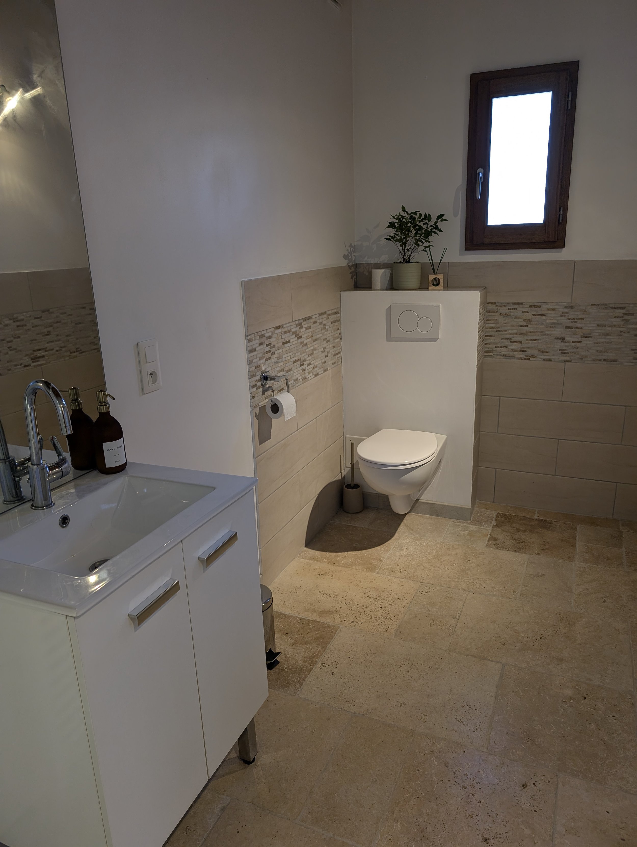 Bathroom with beige tiled floor, a small white sink with soap dispensers, a toilet with a plant on the back ledge, and a window above the toilet.