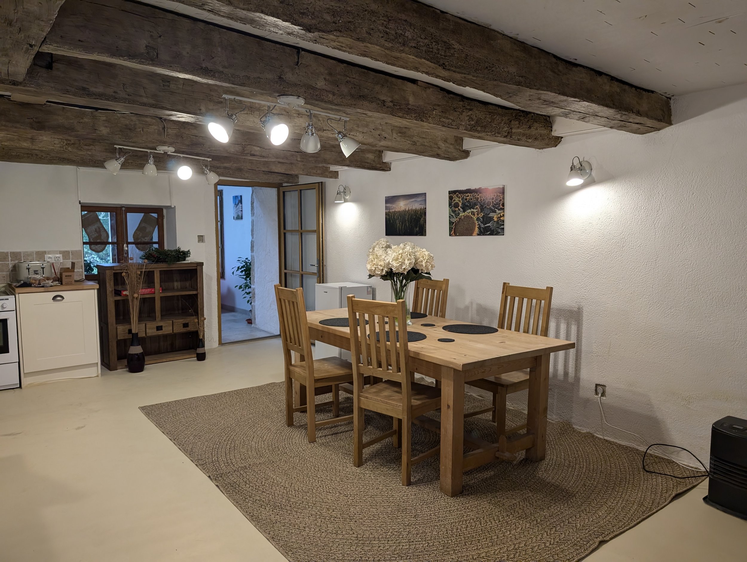 A dining room with a wooden table and six chairs, a vase with white flowers, framed landscape photos on the wall, and a kitchen area with white cabinets and a small window
