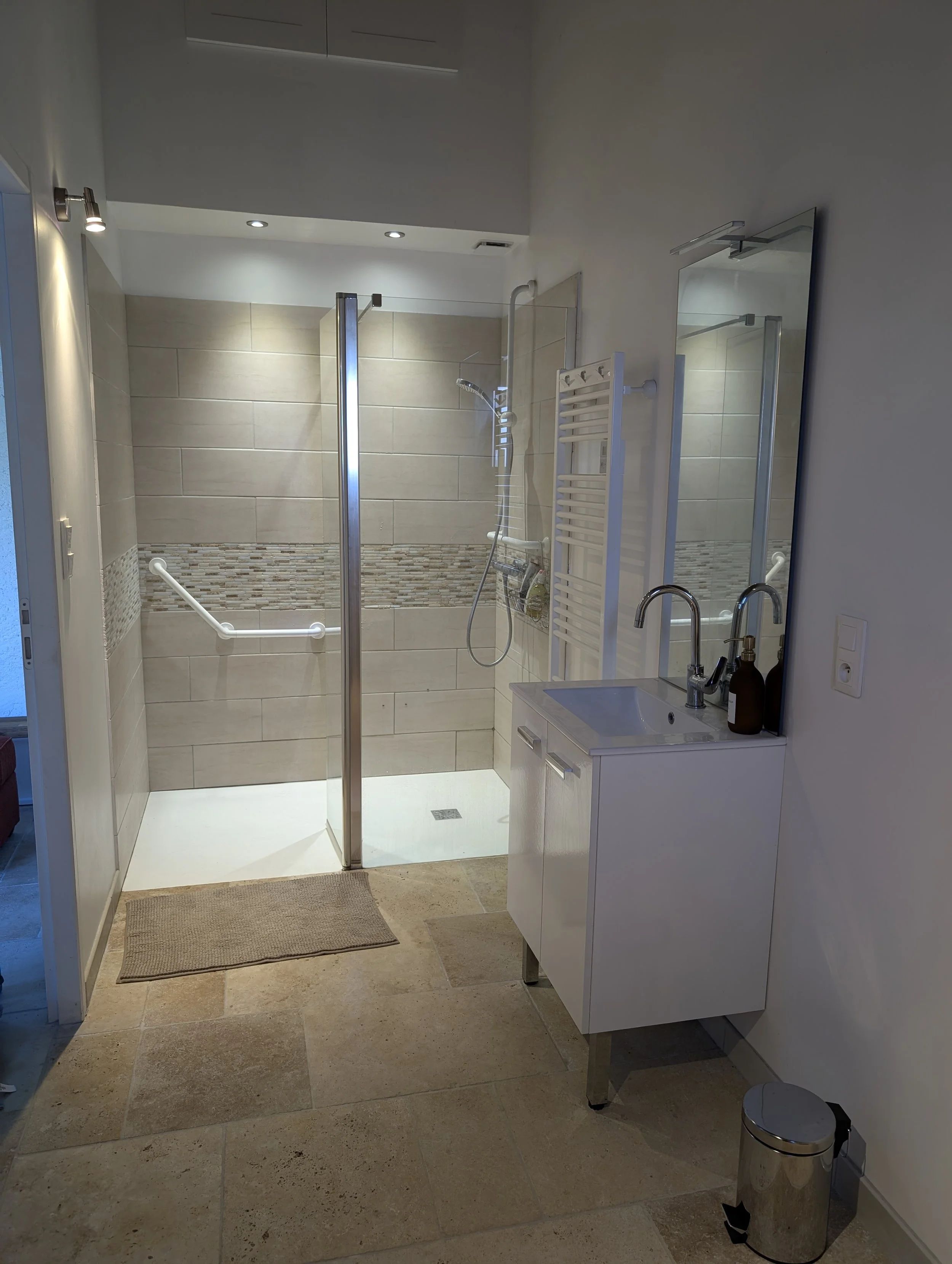 Bathroom with walk-in shower, white vanity with sink, large mirror, beige tiled floor, and wall-mounted radiator.
