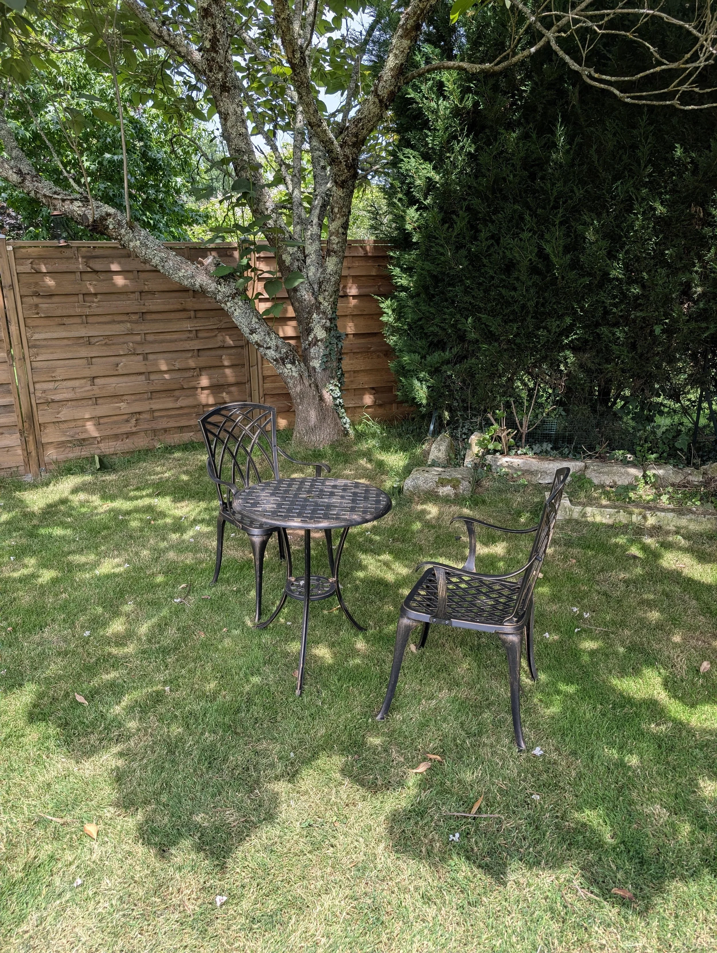 A backyard with a metal table and two black plastic chairs on green grass, surrounded by trees and a wooden fence.
