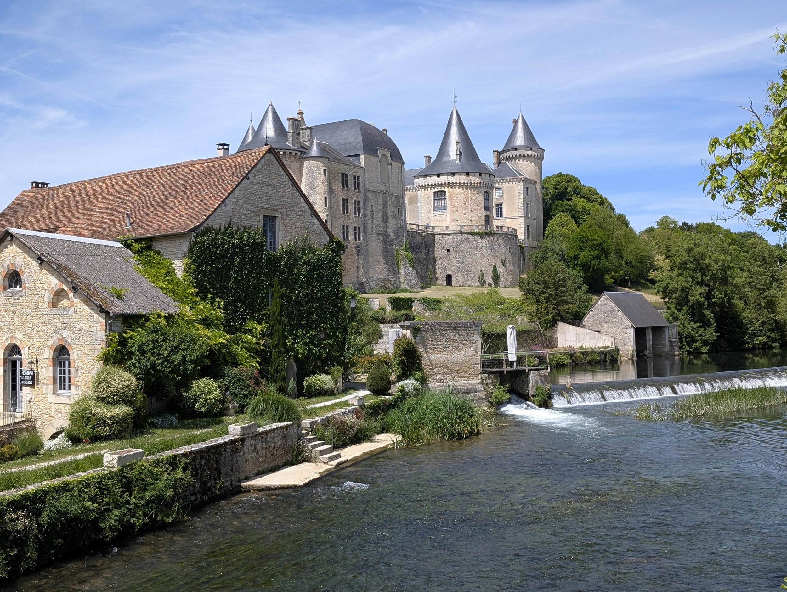 A scenic view of a historic stone castle with turrets, surrounded by lush greenery, beside a flowing river with a small waterfall.