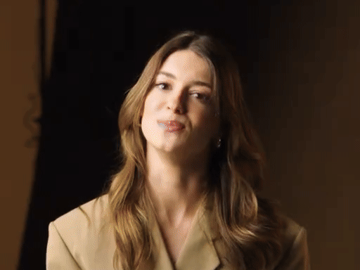 A woman with long, wavy brown hair wearing a beige blazer, looking at the camera with a slight smile.