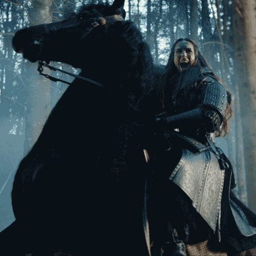 A woman dressed as a medieval knight riding a black horse in a forest.