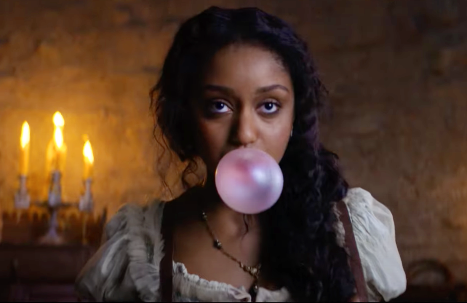 A woman with curly black hair blowing a pink bubble gum bubble, standing in a dimly lit room with a candelabrum with three lit candles in the background.