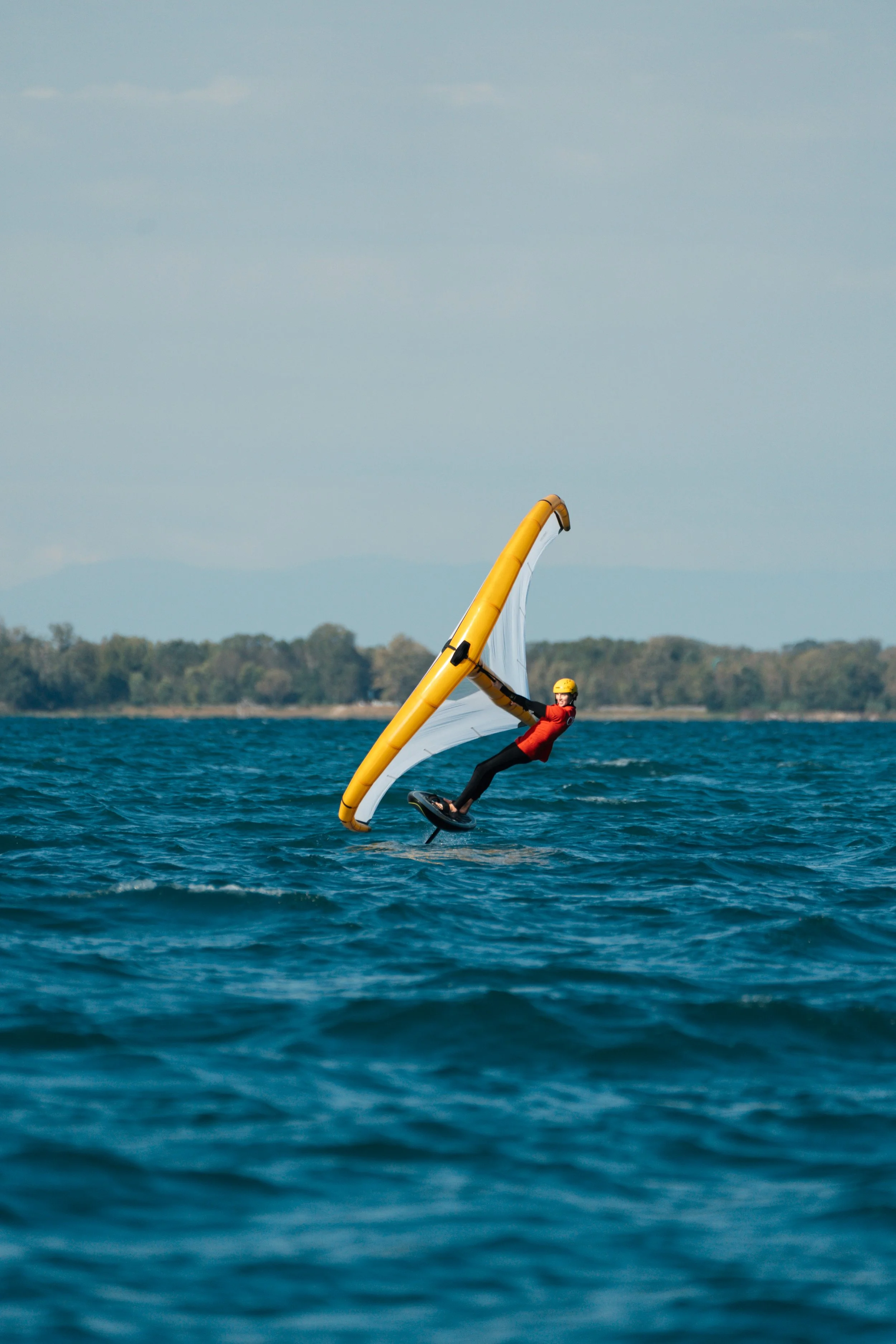 Person windsurfing on the water with a yellow sail and a helmet, with a distant shoreline and cloudy sky in the background.