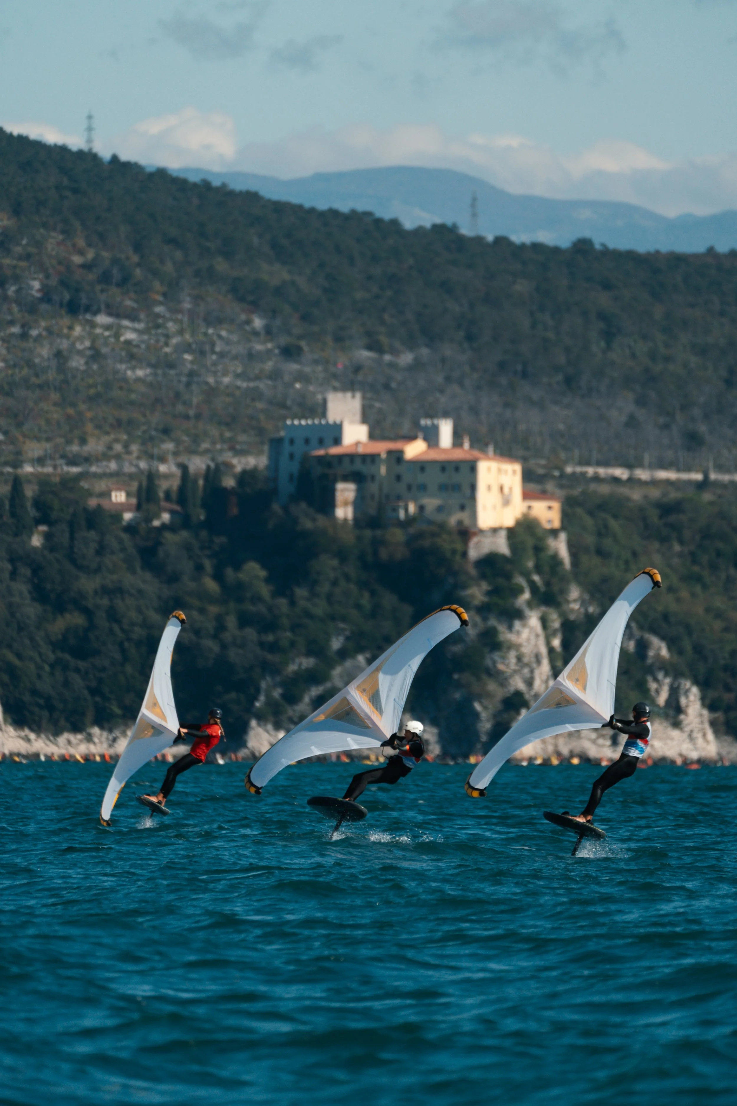 Four people are wakeboarding on a body of water with land and hills in the background. Each person is holding a sail that looks like a curved sheet of fabric, and they are all wearing helmets and life jackets.
