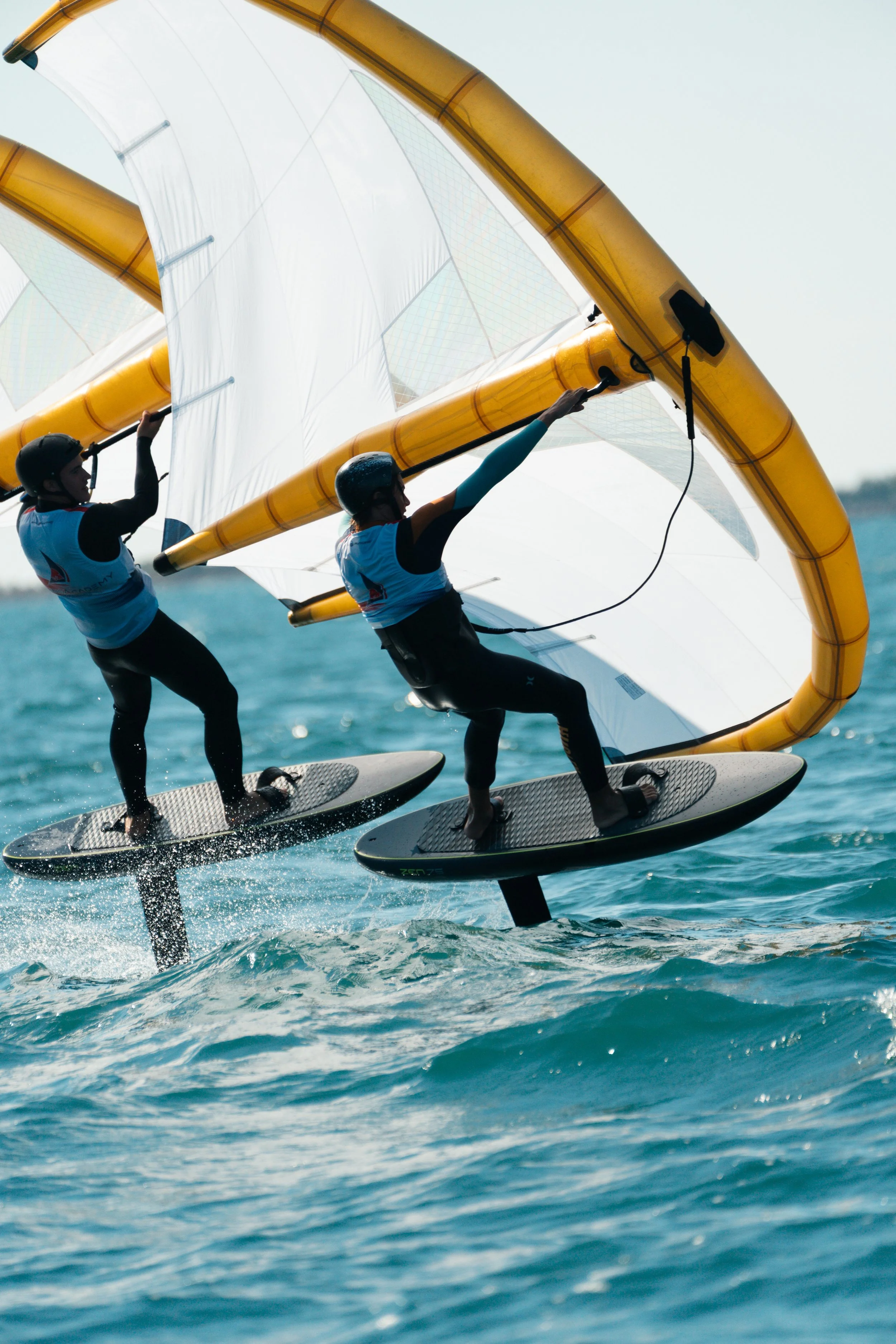 Two people are racing on hydrofoil sailboats on the water, wearing helmets, with large yellow and white sails above them.