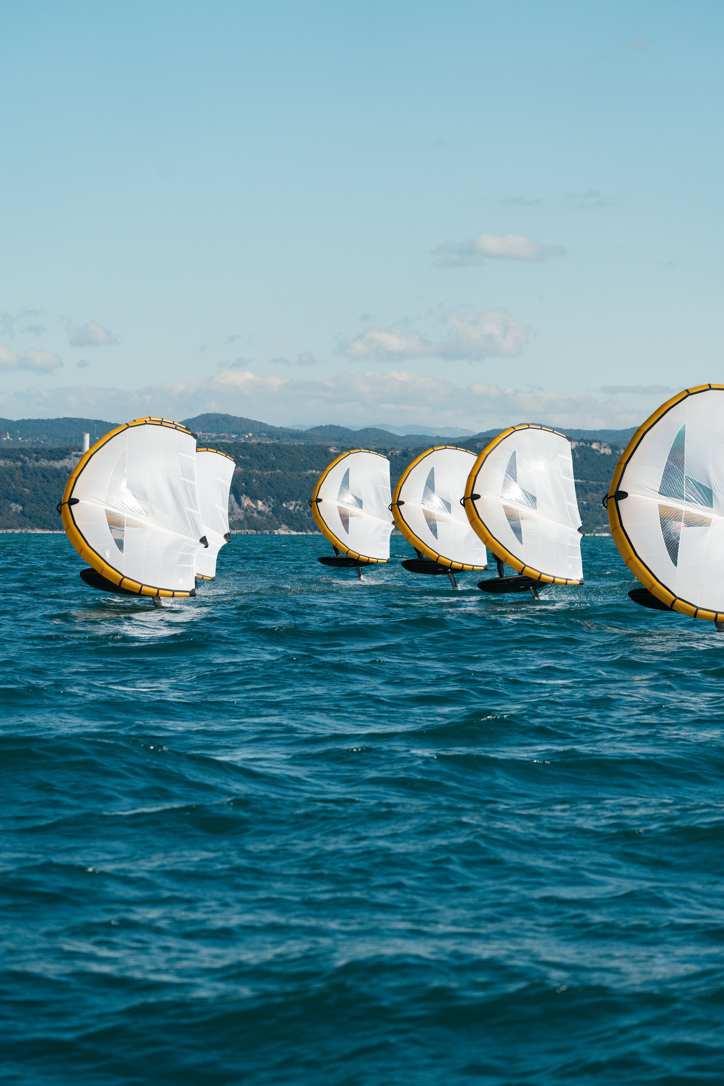 A fleet of windsurfing sails on the water, with mountain hills in the background under a partly cloudy sky.