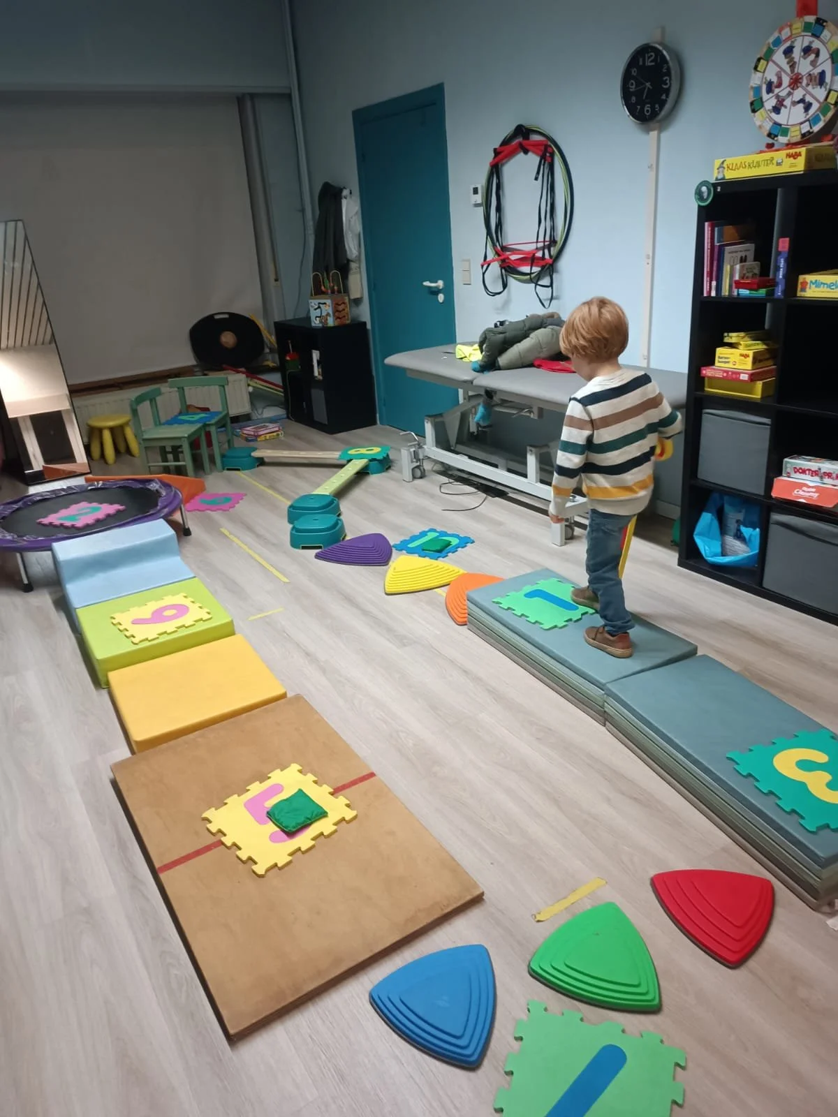 Indoor playroom set up with foam mats, balance beam, children’s toys, and a boy walking on foam stepping stones.