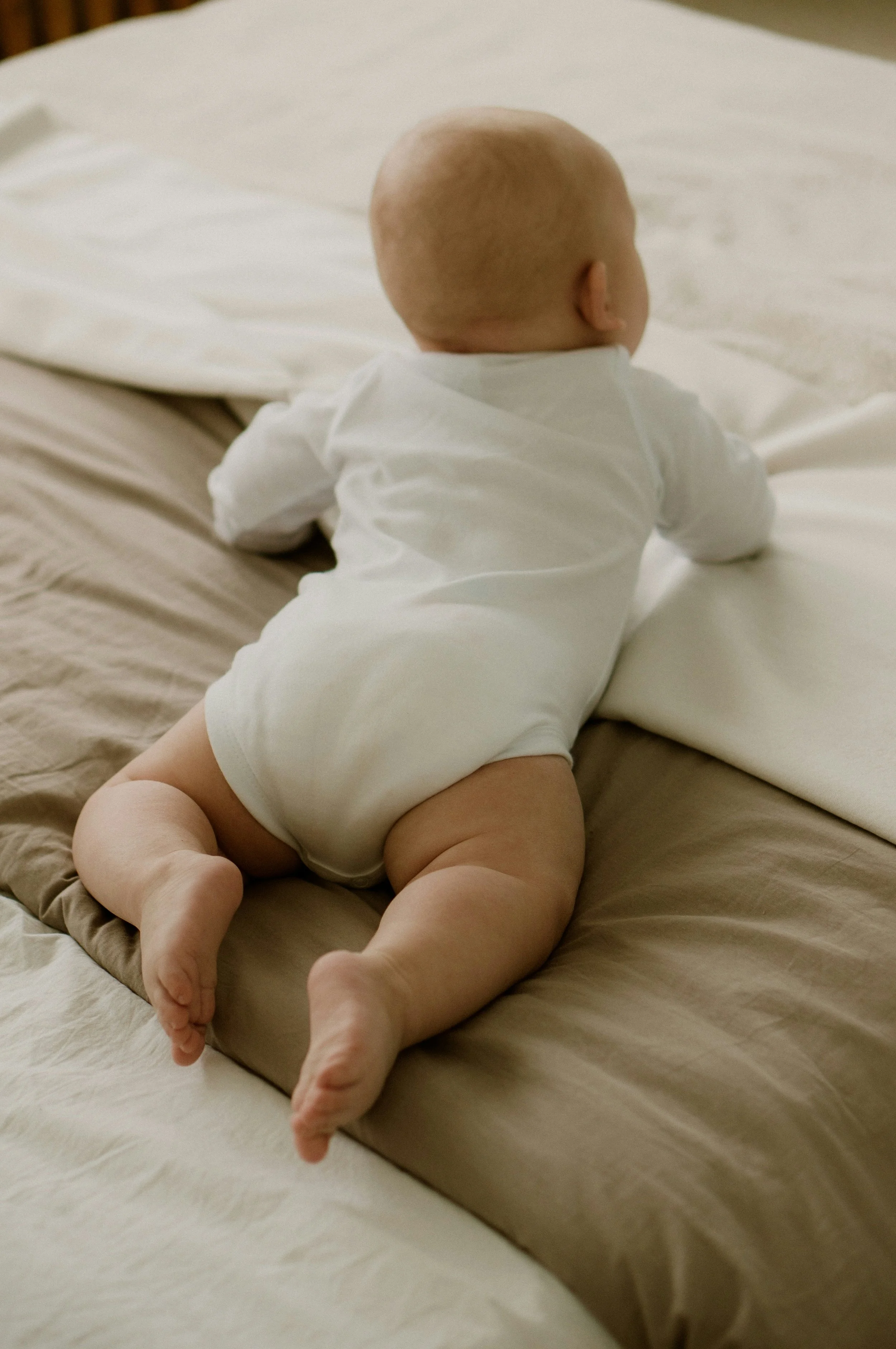 A baby crawling on a bed, facing away, wearing a white onesie with a beige blanket underneath.