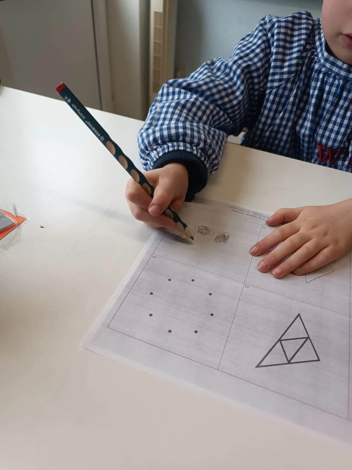 A child wearing a blue checkered shirt drawing on a worksheet with a colored pencil.