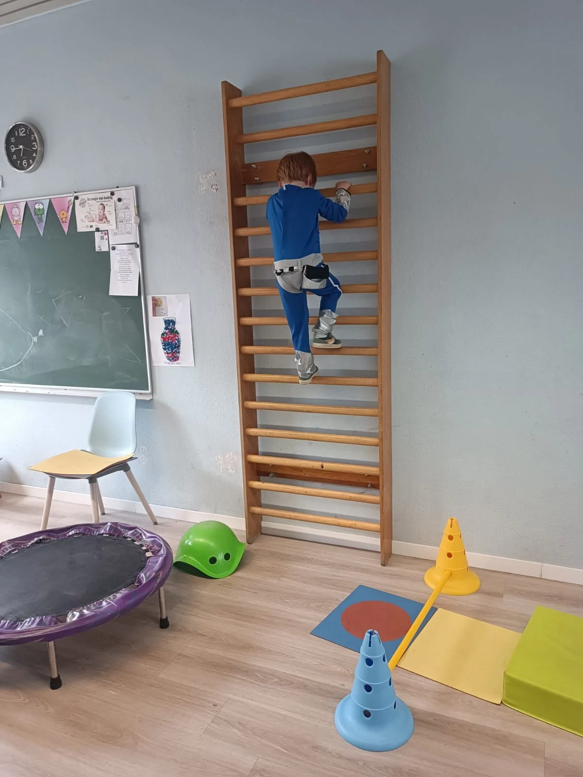 Child climbing a wooden wall ladder in a playroom, with a trampoline, traffic cones, and yarn mats on the floor, and a chalkboard on the wall.