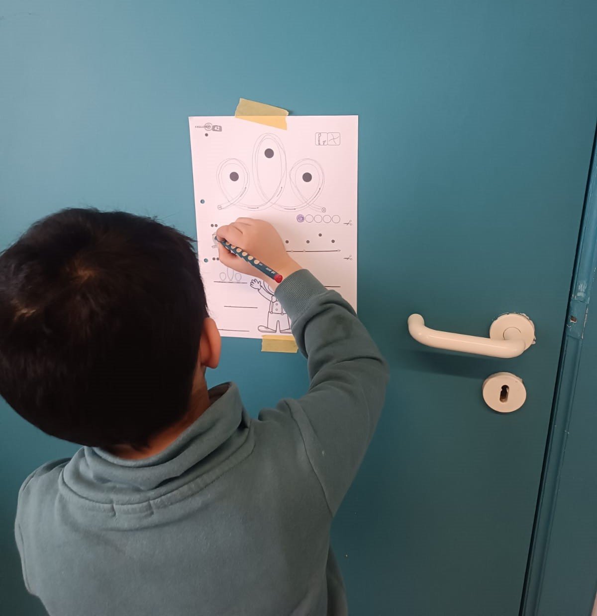 A young boy with short black hair drawing on a worksheet taped to a blue wall. The worksheet has illustrations and dots for connecting patterns. The boy is holding a black and red pencil.