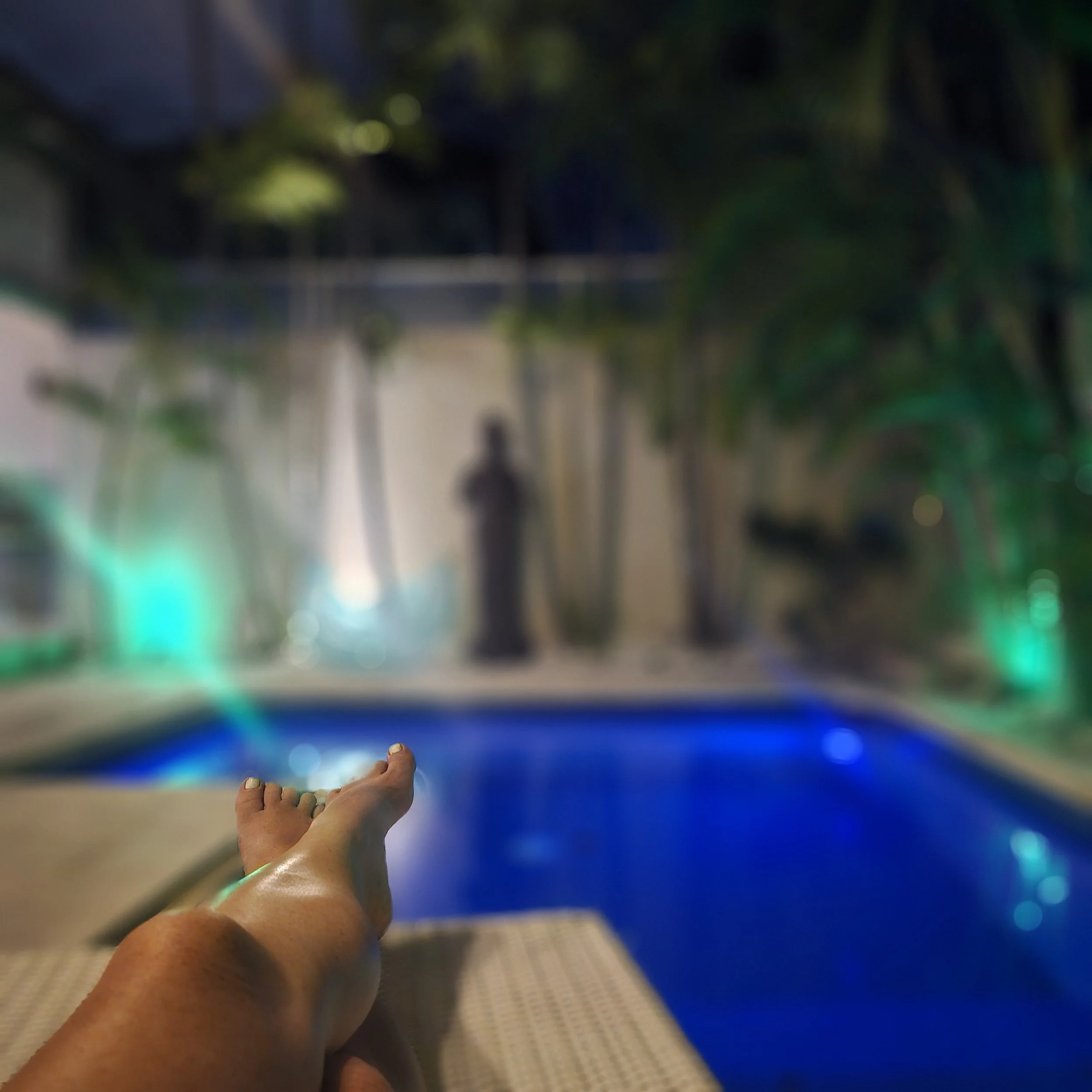 A person relaxing by a swimming pool at night with their legs extended on a lounge chair, illuminated by colorful pool lights and surrounded by lush greenery.