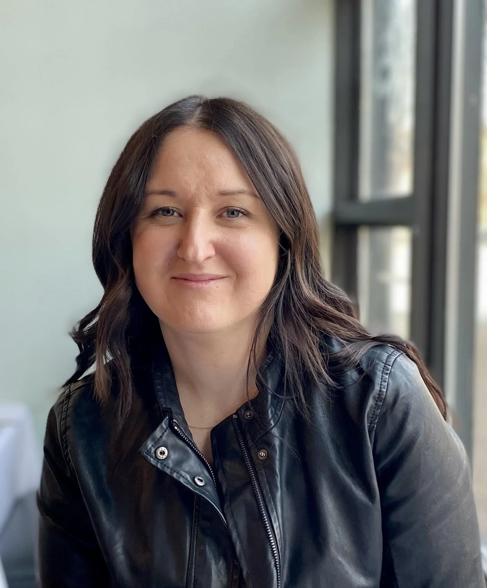 a woman with long dark hair and blue eyes wearing a leather jacket smiling at camera