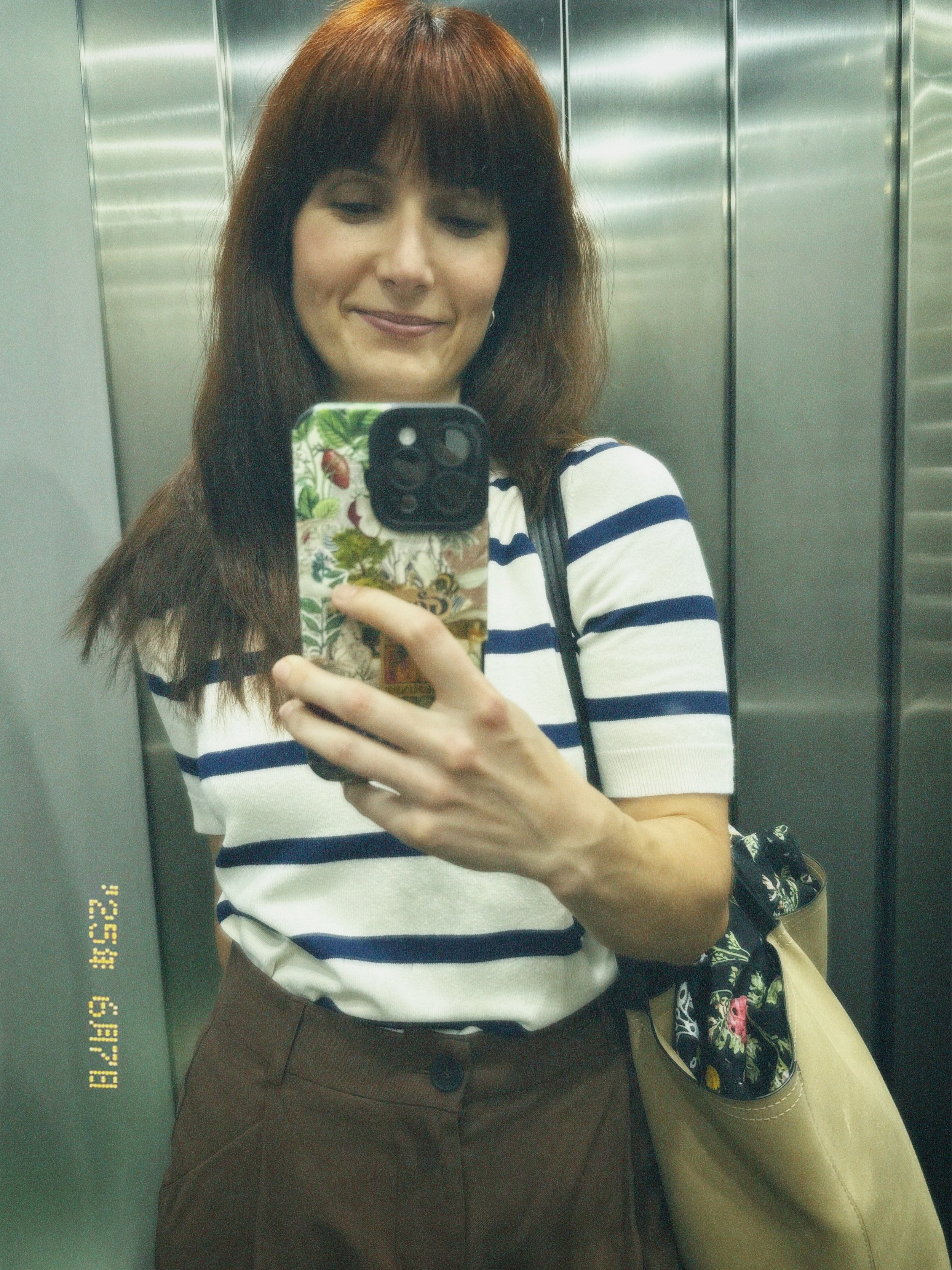 Woman with long brown hair taking a mirror selfie in an elevator, smiling with a striped shirt, brown pants, and carrying a floral bag.