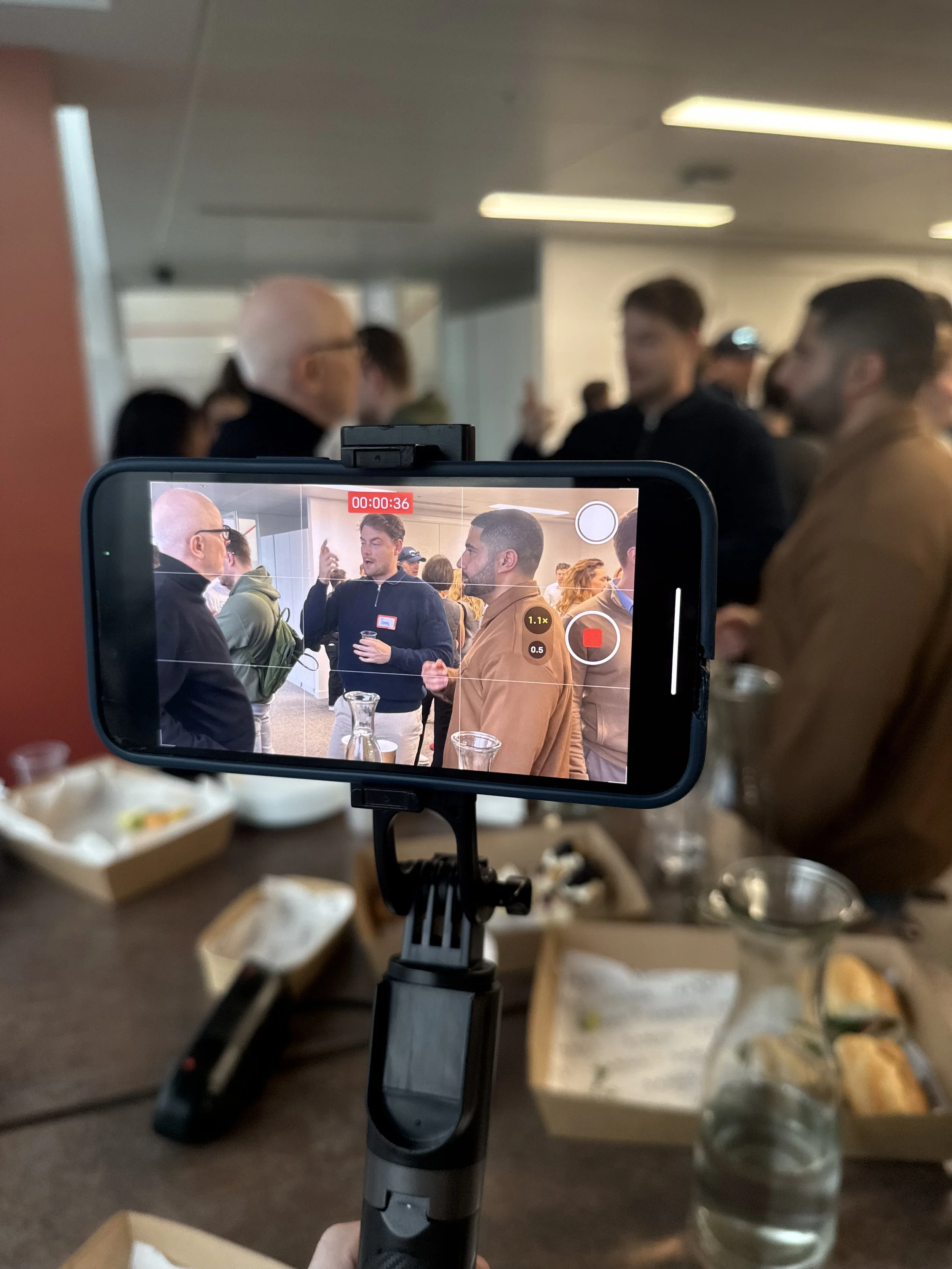 A smartphone on a selfie stick recording a crowded indoor event, showing two men in conversation in the background, with food and drink on the table in the foreground.