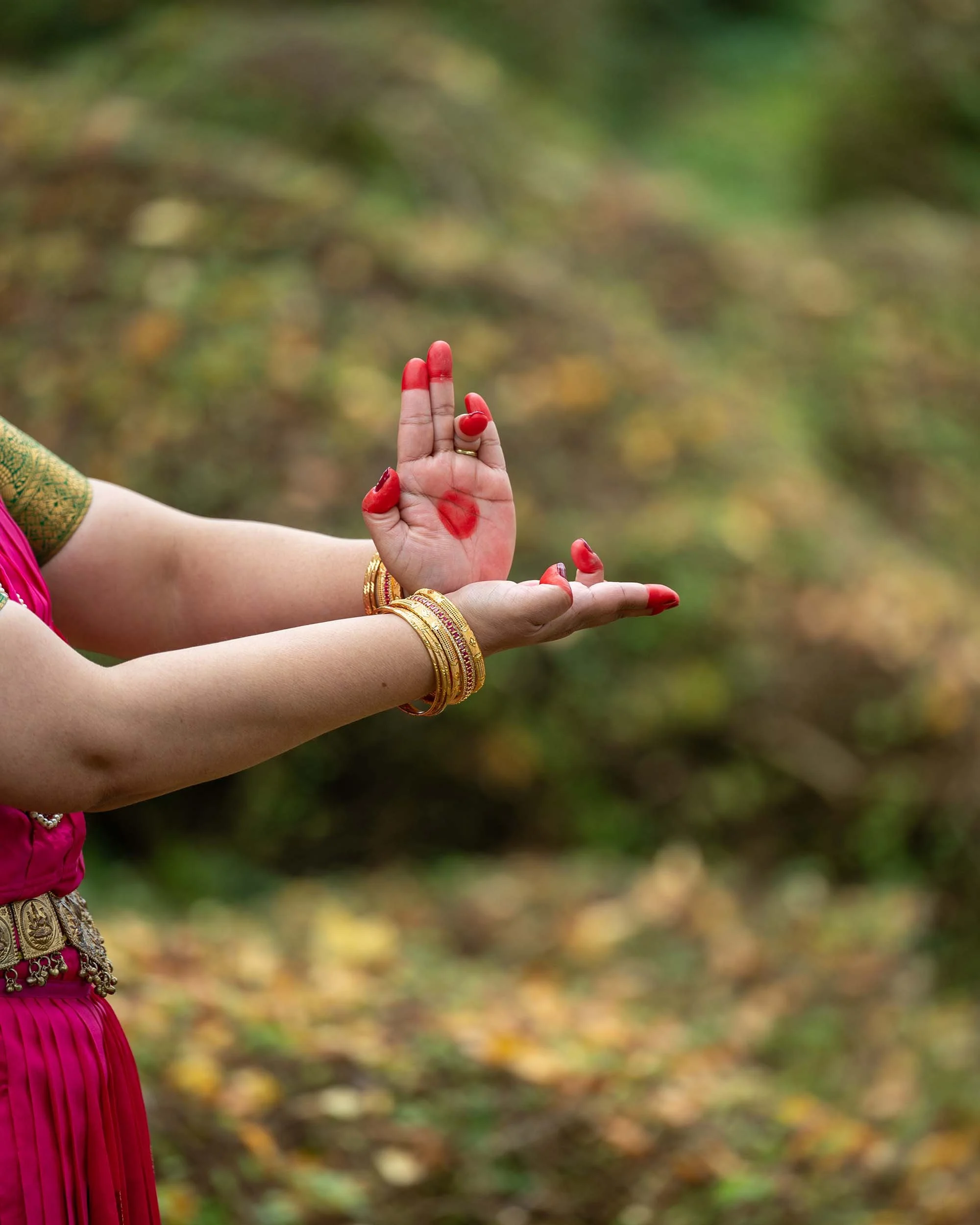 A person wearing traditional Indian clothing and jewelry performing a mudra with their hands in an outdoor setting with blurred autumn foliage in the background.