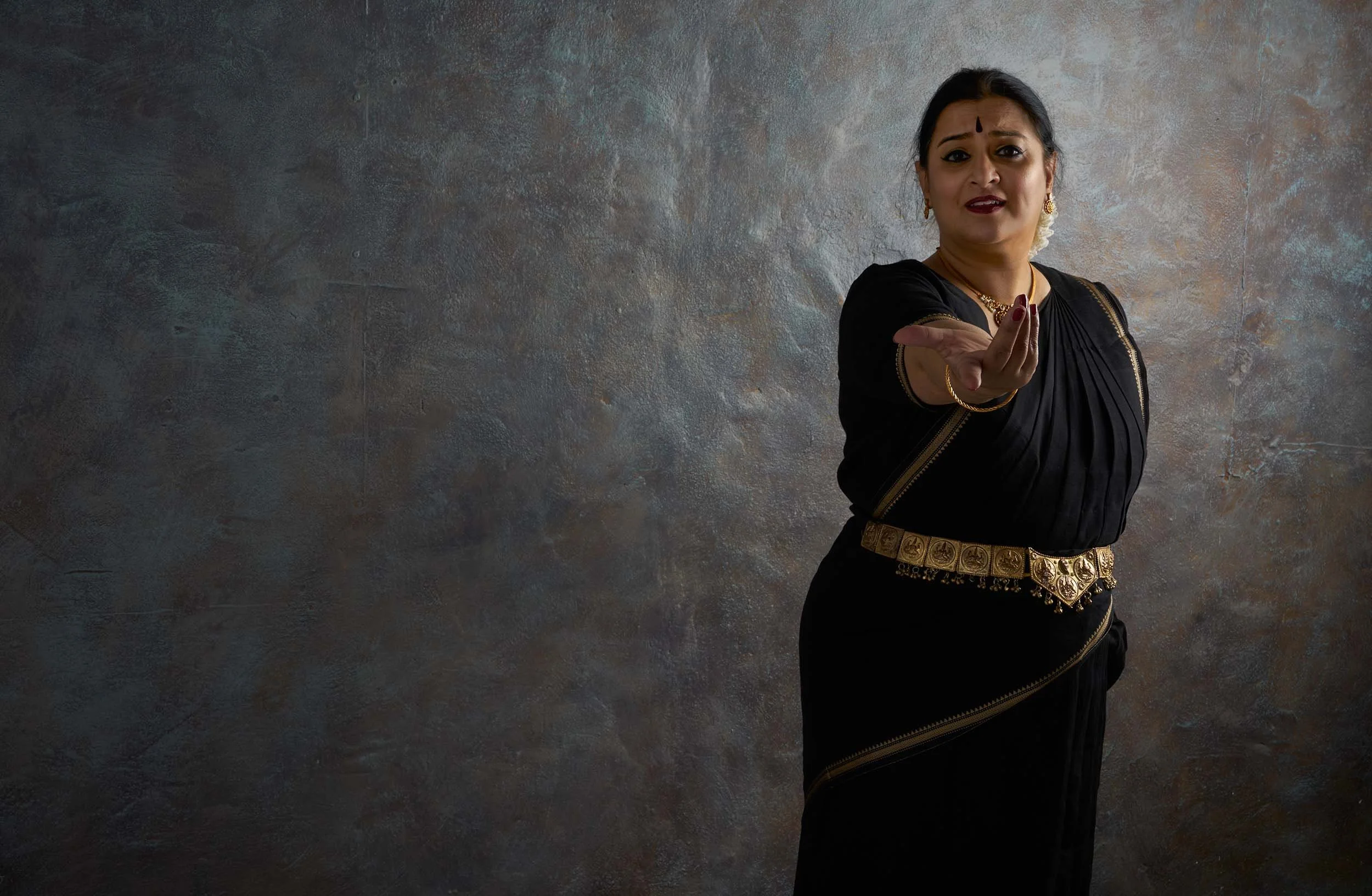 A woman dressed in traditional Indian attire, reaching out with her hand, set against a textured gray background.