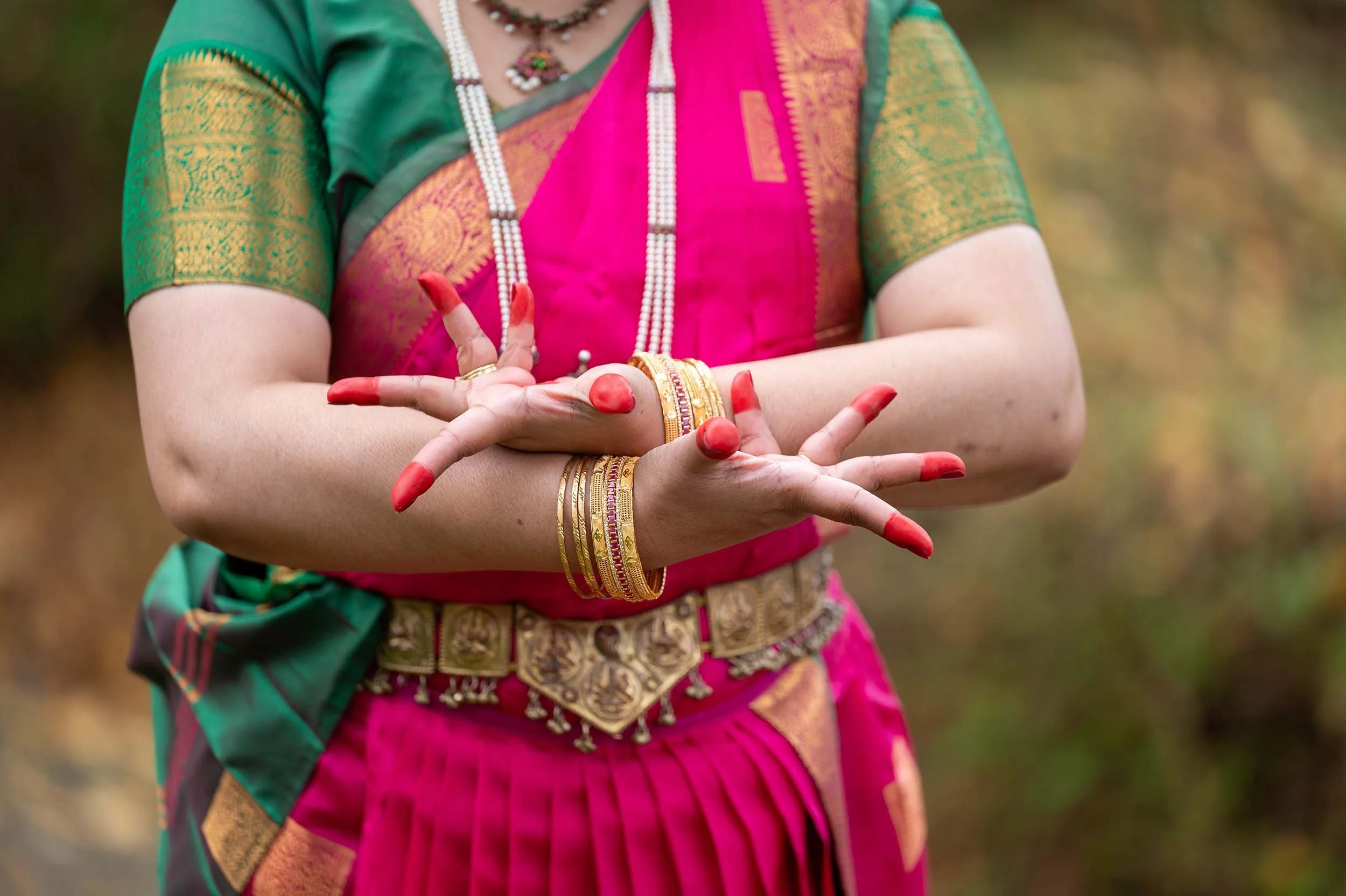 Bharatanatyam dancer's hands in mudra pose wearing gold bangles, red nails, and pink silk saree.