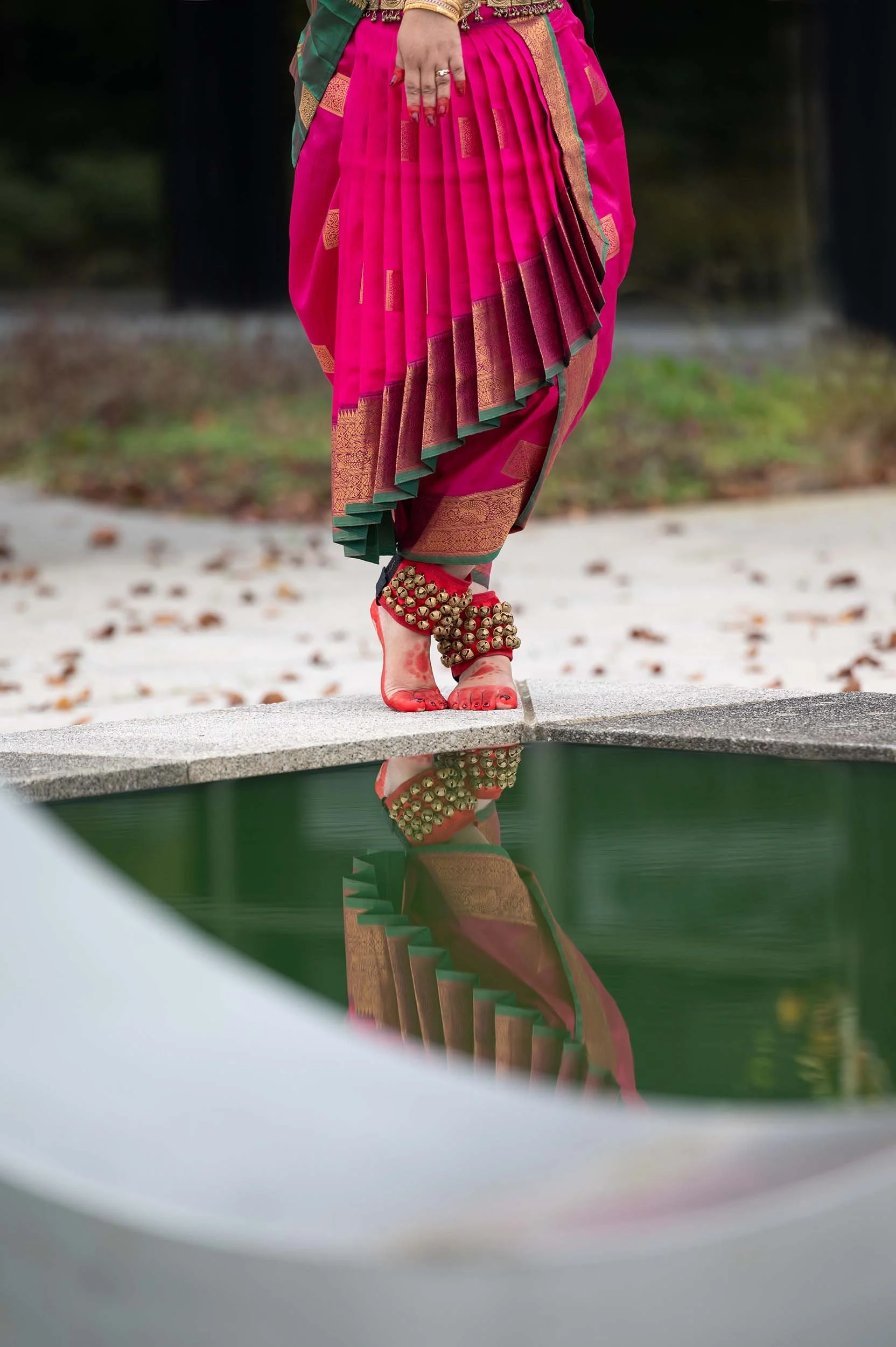 A person dressed in traditional Indian attire, wearing a pink and gold saree with pleats, standing near a reflective pool of water. The person has decorated anklets with bells and is standing barefoot, with their reflection visible in the water.