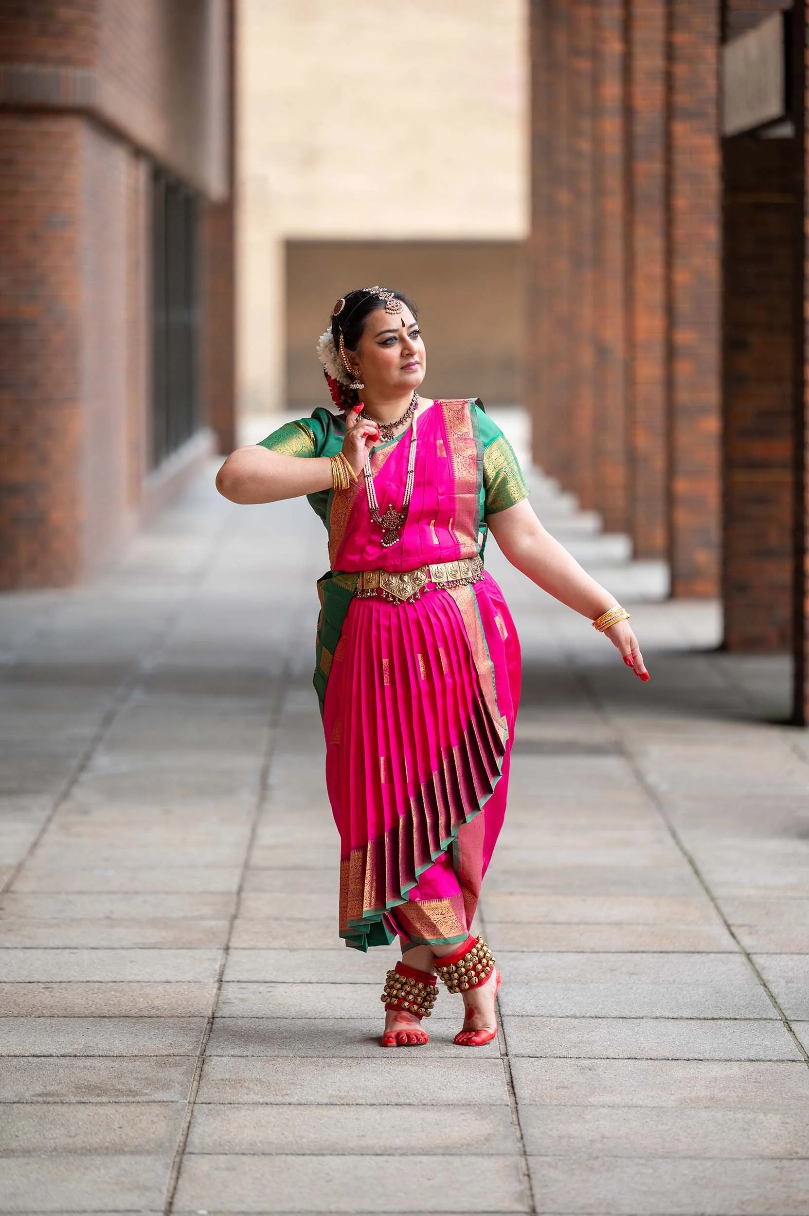 Bharatanatyam dancer in a pink and green costume posing outdoors between brick columns, wearing traditional jewellery and ankle bells.