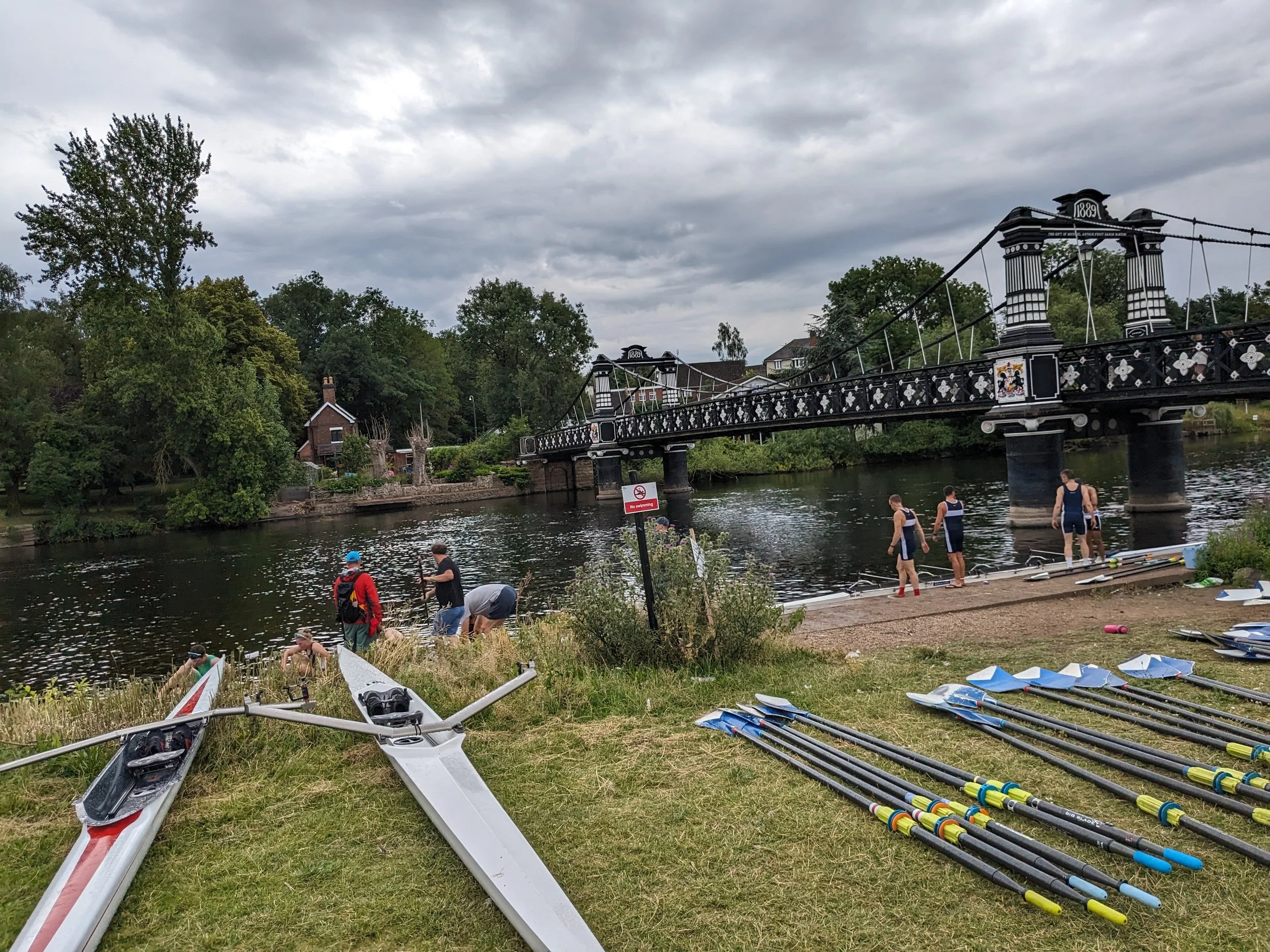 People preparing and launching rowing shells by a river, with oars laid out on the grass and a decorative iron bridge in the background under cloudy skies.