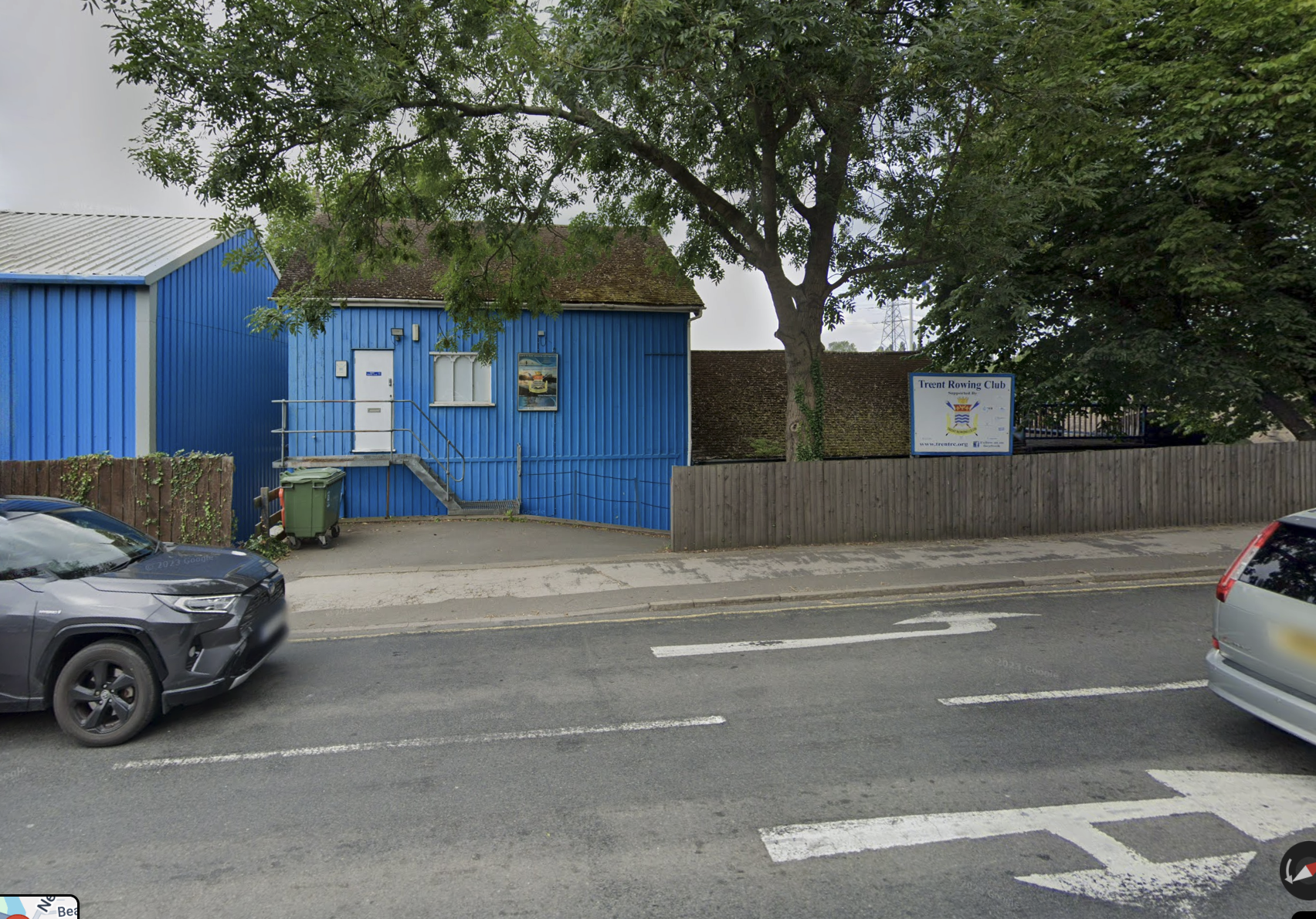 Street view with parked cars, blue building with stairs, tree, and sign for Trent Rowing Club.